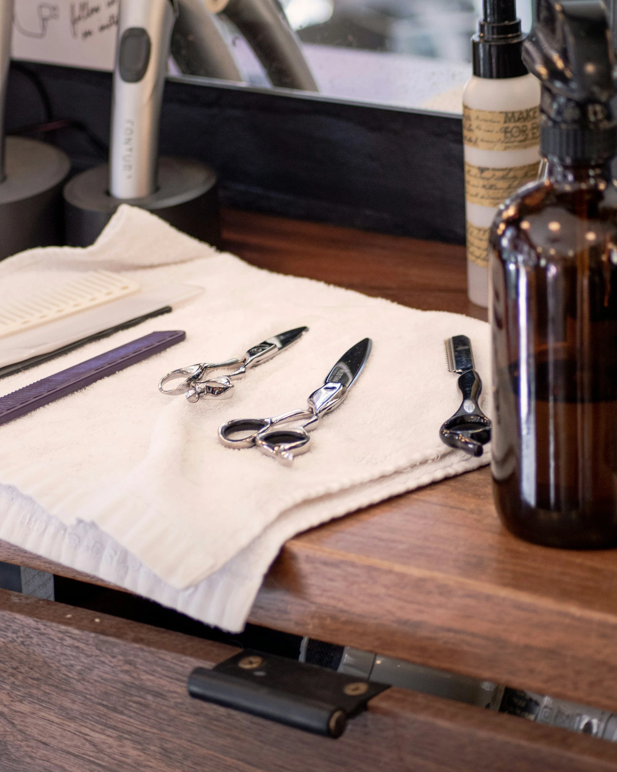 A white towel with several pairs of hair cutting scissors, a straight razor, and a hair clipper laying on it, with various hair care products and tools, on a wooden barber station.