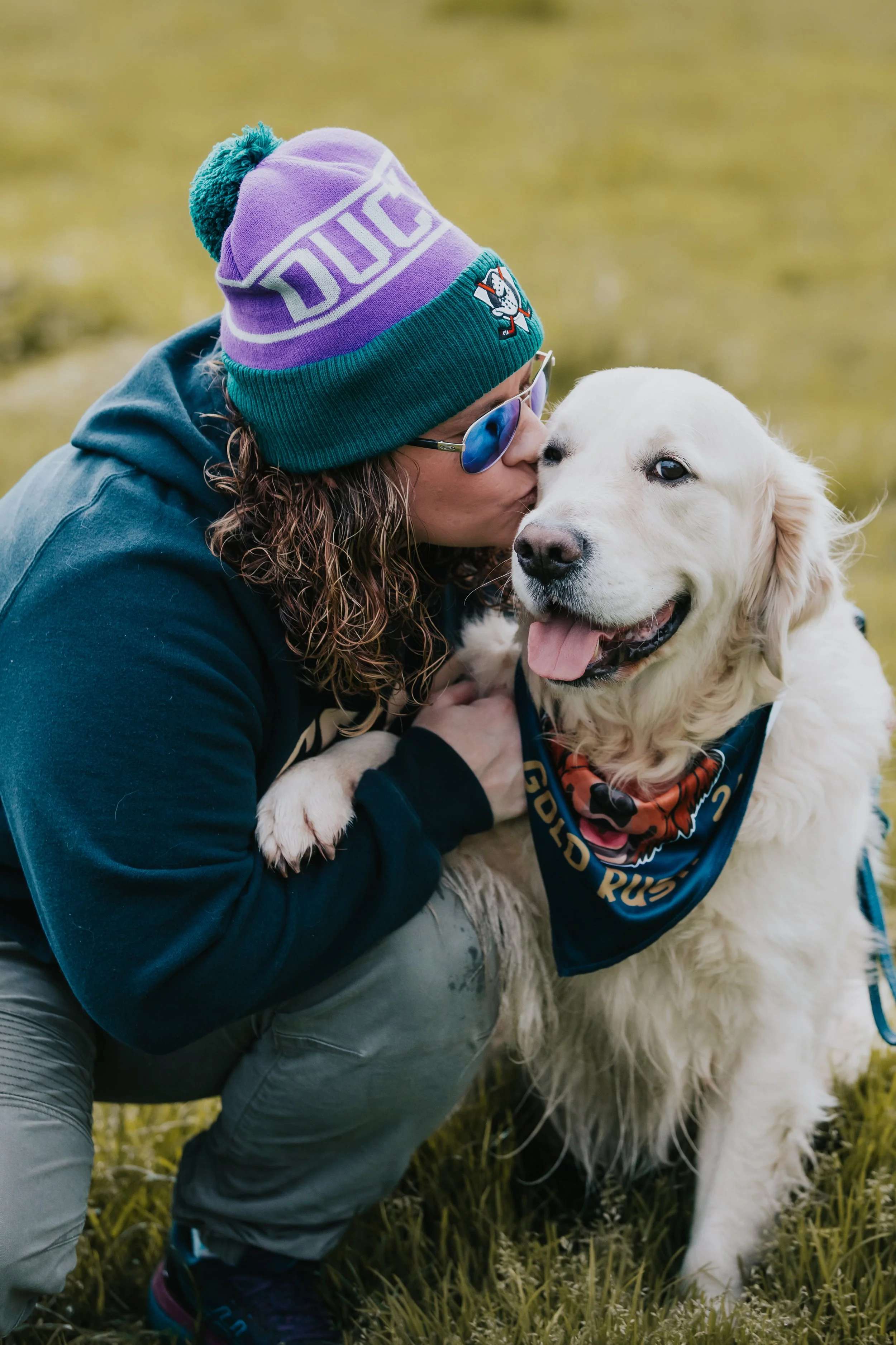 Person wearing a colorful beanie, sunglasses, and a dark hoodie hugging a Golden Retriever wearing a blue bandana, outdoors on grass.