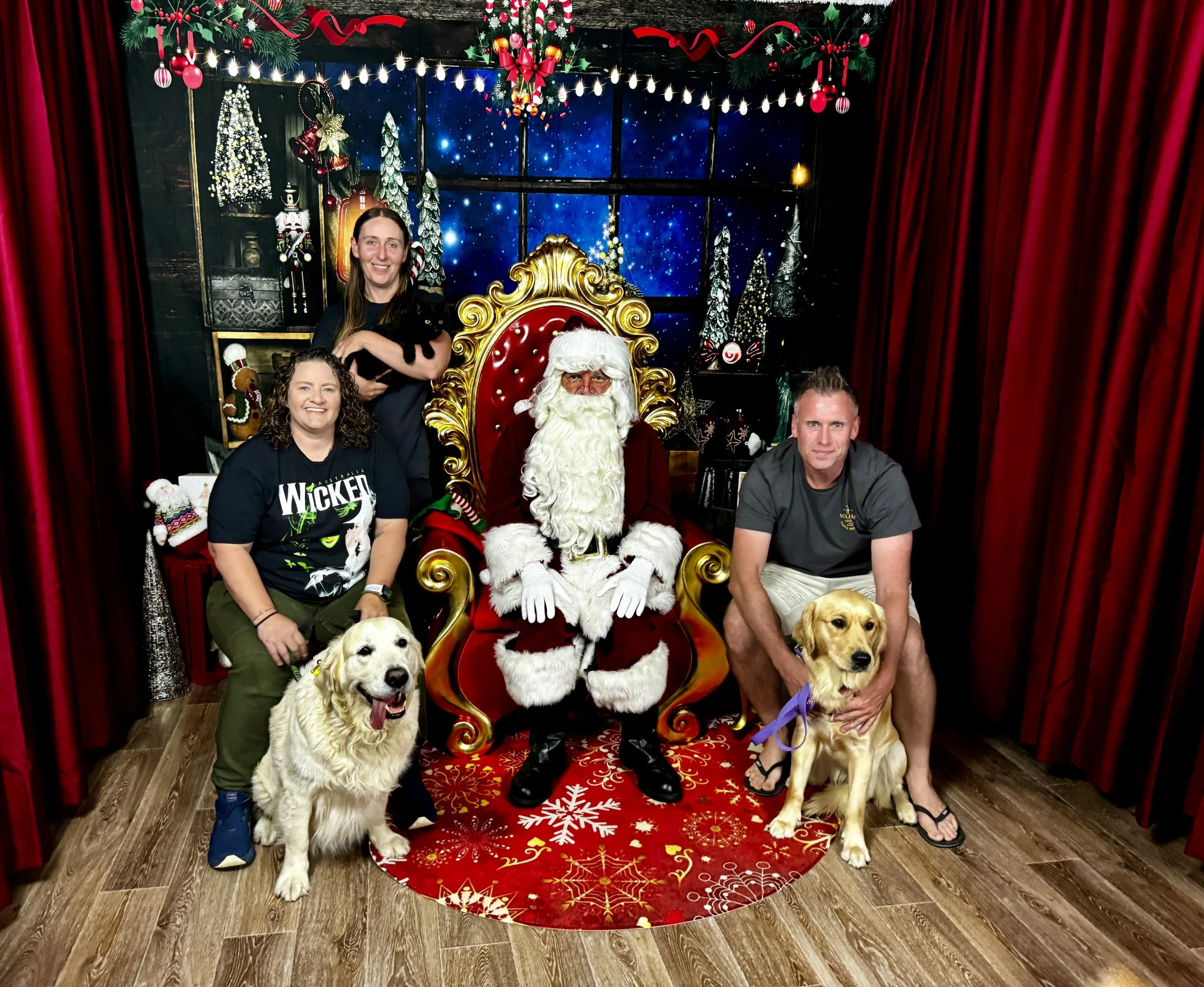 A festive holiday scene with Santa Claus seated on a red and gold throne, flanked by two dogs and surrounded by three smiling people. The background features holiday decorations, a snowy winter scene, and red curtains.