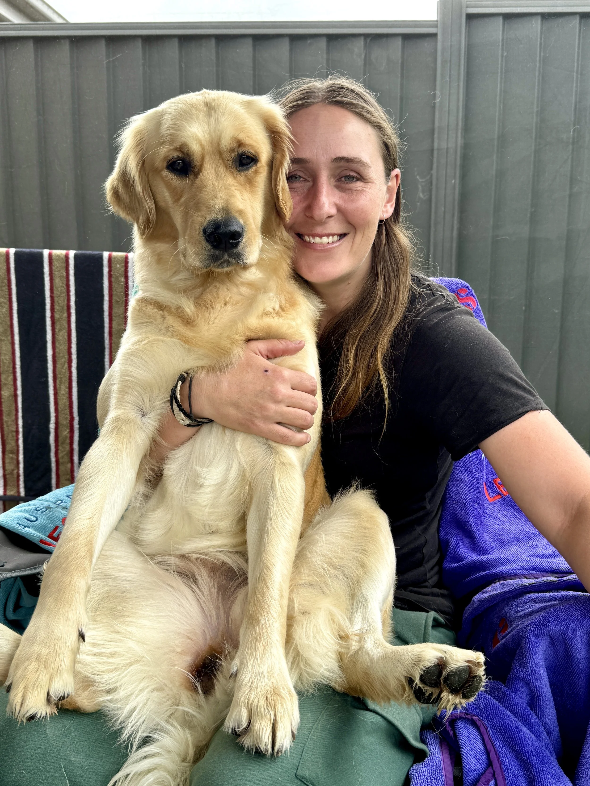 Woman smiling and holding a golden retriever puppy in an outdoor setting with a green fence in the background.
