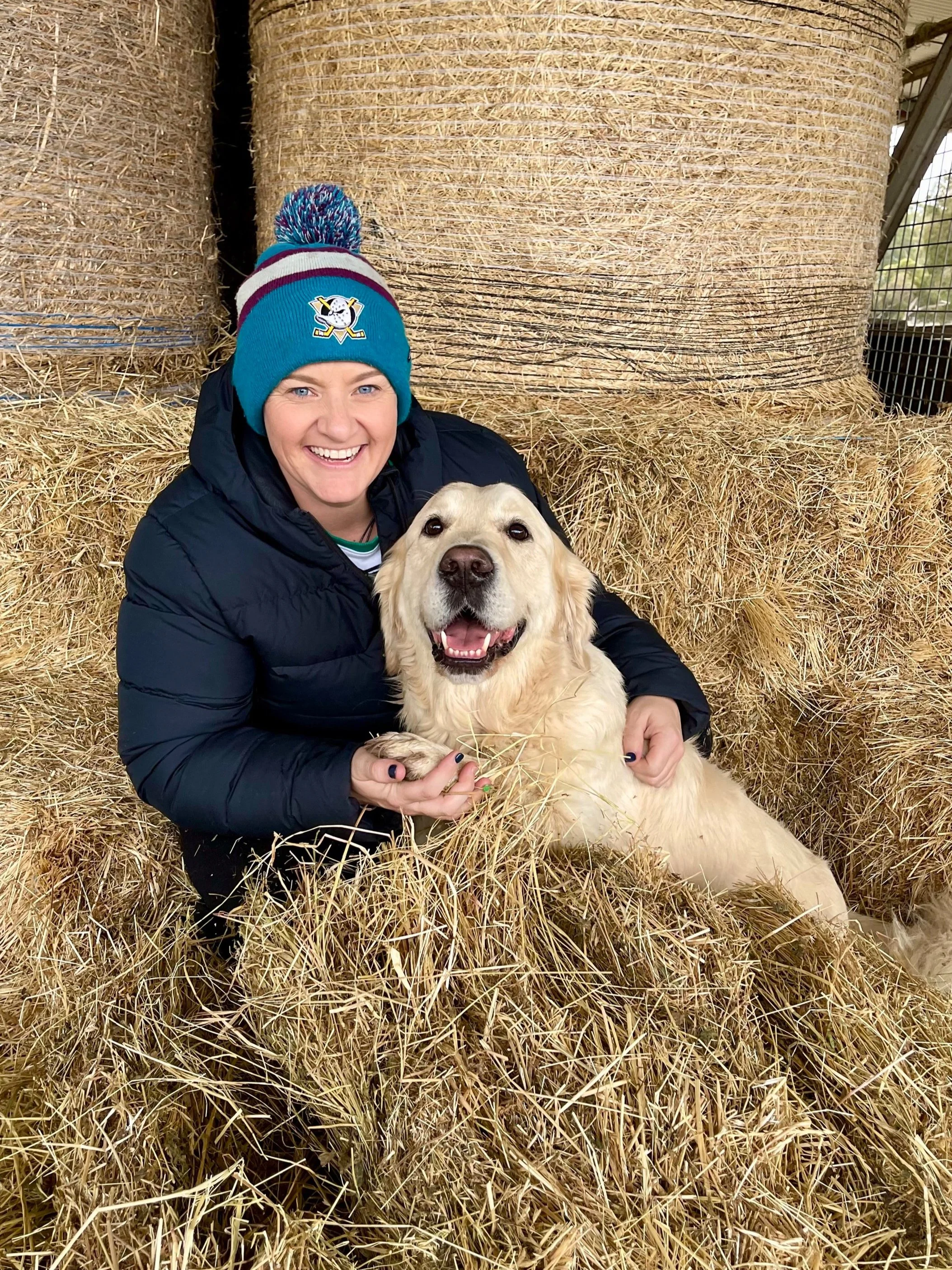 A smiling woman wearing a blue knit hat with a playful logo and a black jacket, holding a happy golden retriever puppy amidst hay bails indoors.