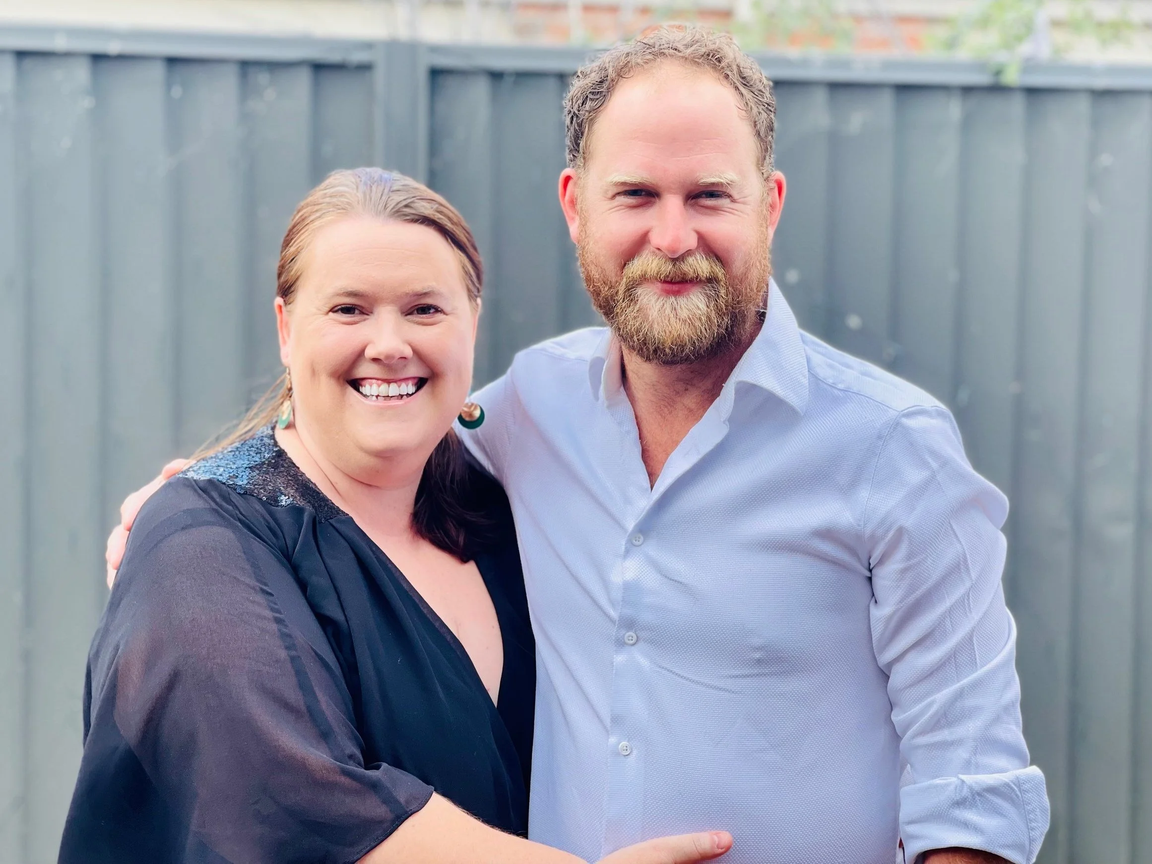 A smiling woman and man with light skin standing close together outdoors, in front of a wooden fence. The woman has dark hair and is wearing a black top, and the man has a beard, short hair, and is dressed in a light blue shirt.