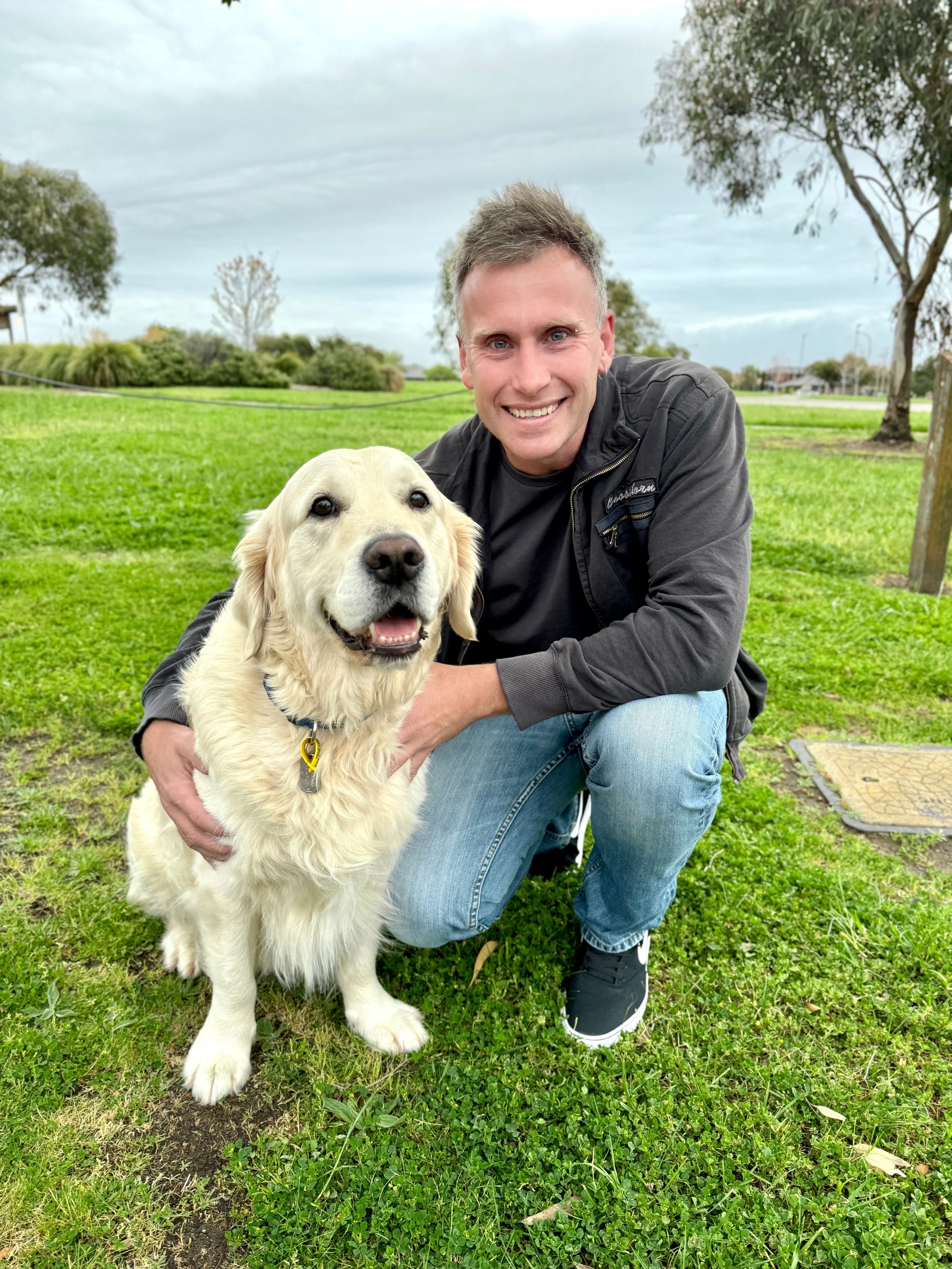 A man kneeling on the grass, smiling, with a golden retriever dog sitting beside him outdoors in a park with trees and cloudy sky.