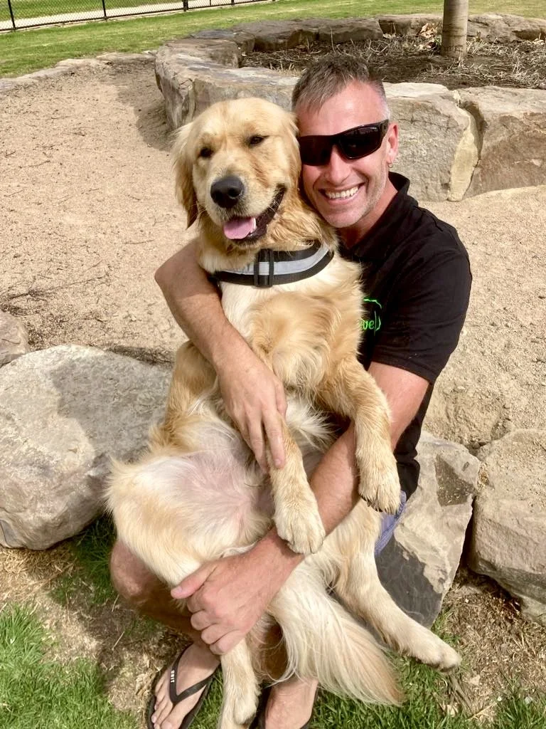 A man wearing sunglasses and a black shirt smiling and hugging a large golden retriever dog outdoors in a park with rocks and grass.
