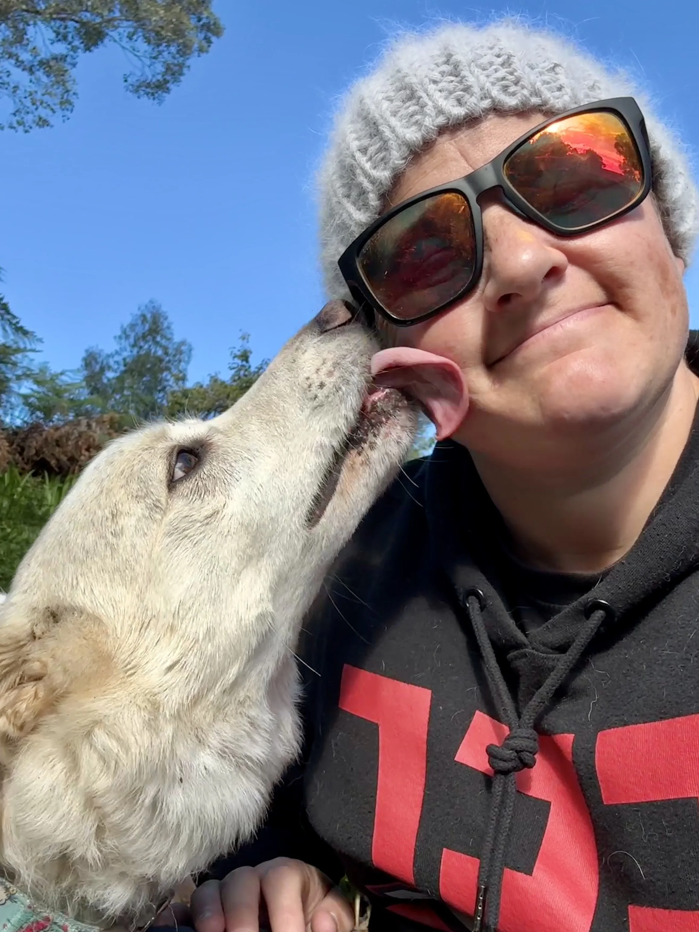 A woman in sunglasses and a gray hat is smiling as a young, light-colored puppy licks her face.