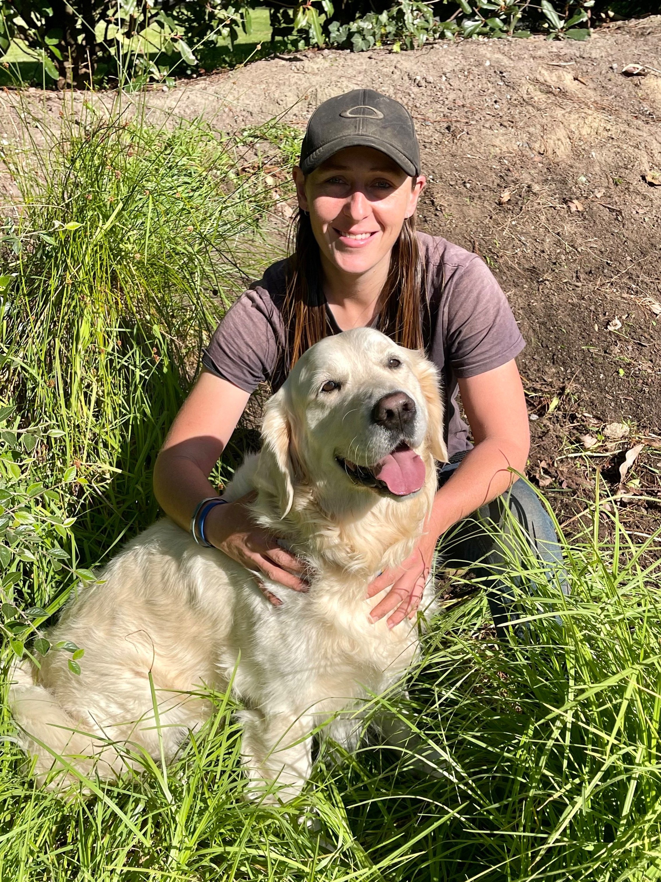 A woman with long brown hair, wearing a black cap and a gray t-shirt, sitting outdoors on dirt and surrounded by green grass and plants, smiling with a golden-colored Labrador Retriever dog, also smiling and with its tongue out.