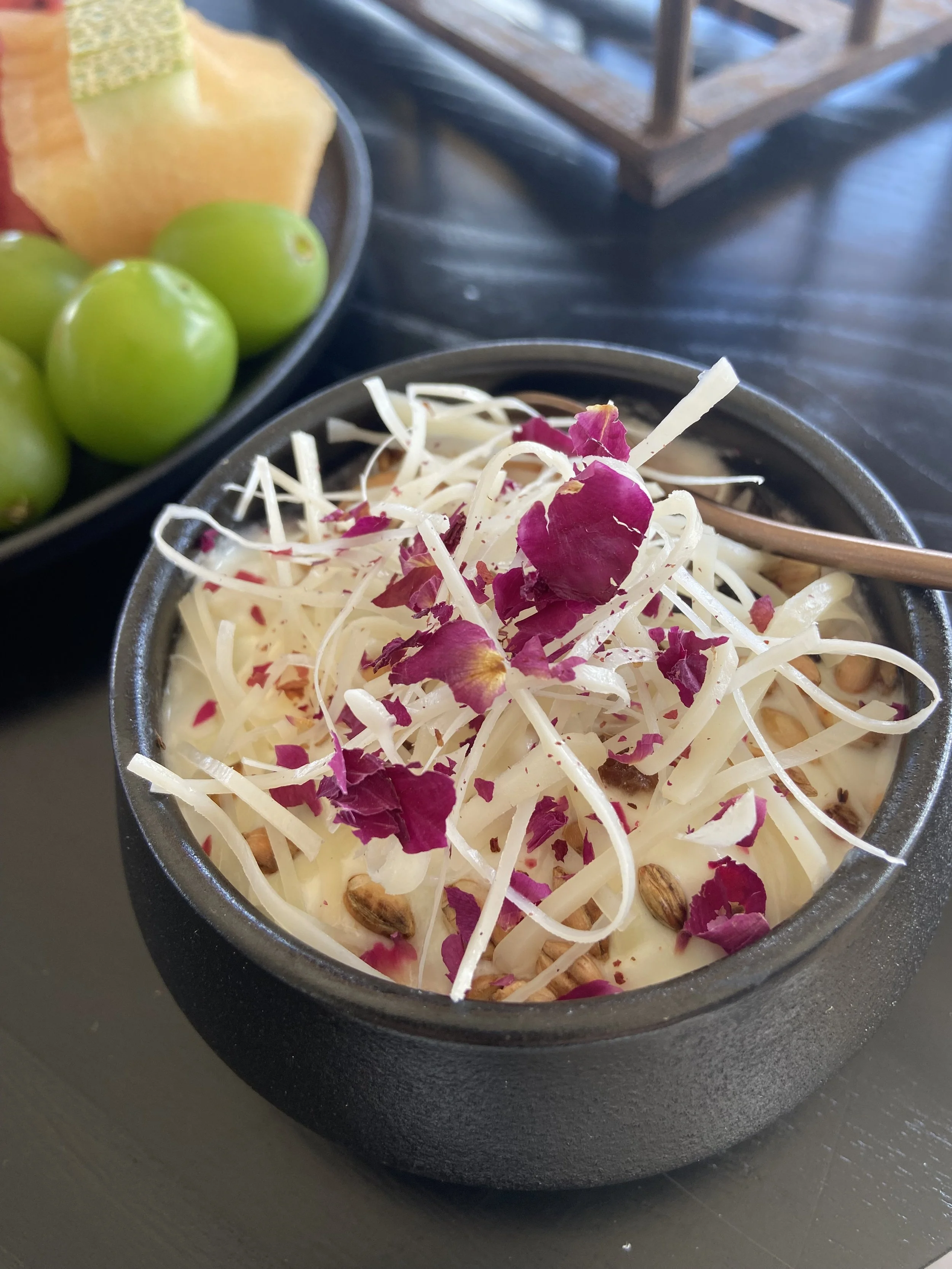 A black bowl filled with shredded cheese, rose petal bits, and seeds, likely a cheese or dip dish. In the background, a plate of green grapes and cut melons can be seen.