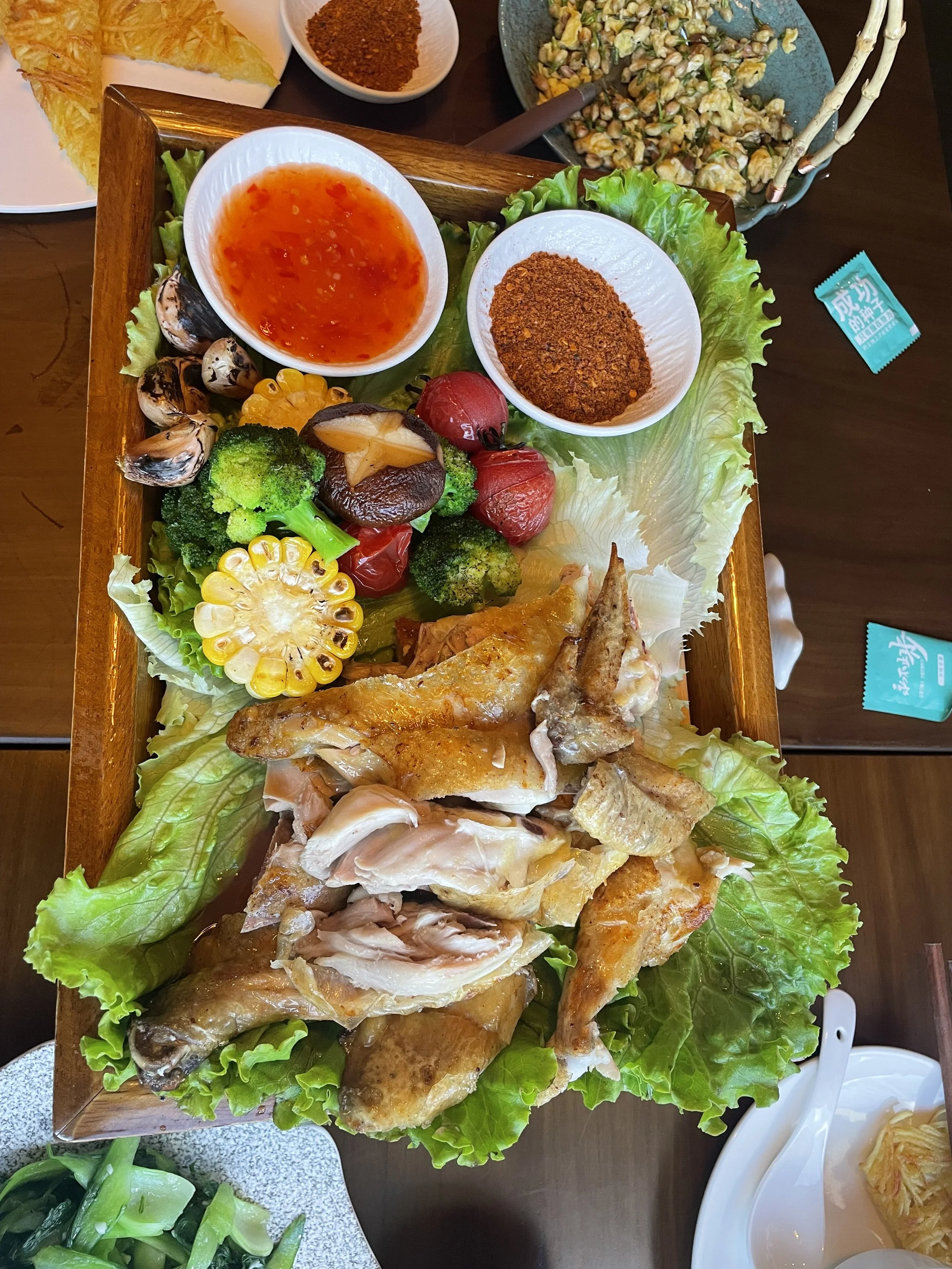 A large wooden platter of roasted chicken with leafy greens, cherry tomatoes, broccoli, corn, mushrooms, and dipping sauces, with side dishes including a vegetable salad and condiments.