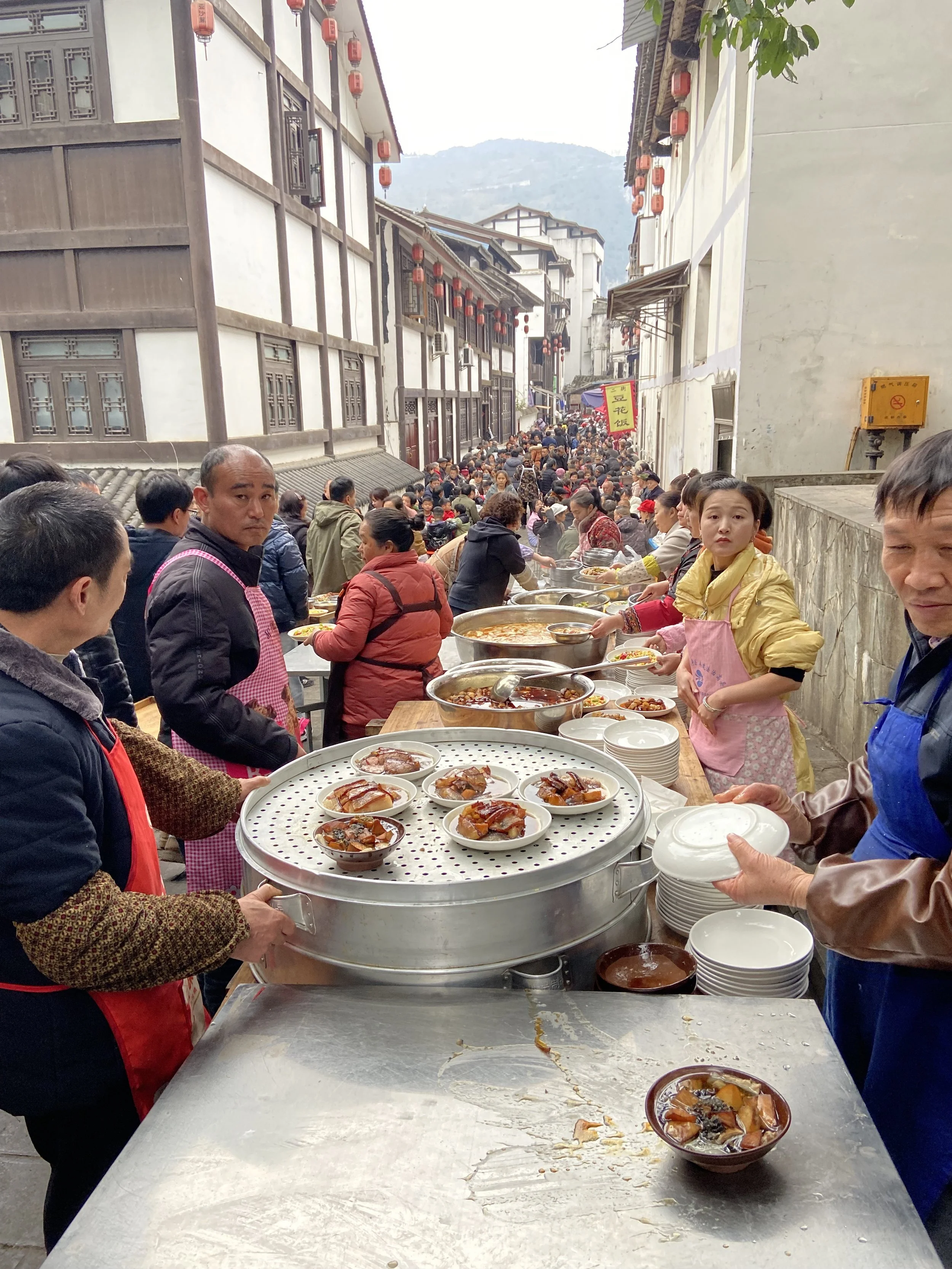 Street food market in a traditional Chinese area with tables of dishes, many people, and narrow streets with lanterns hanging overhead.