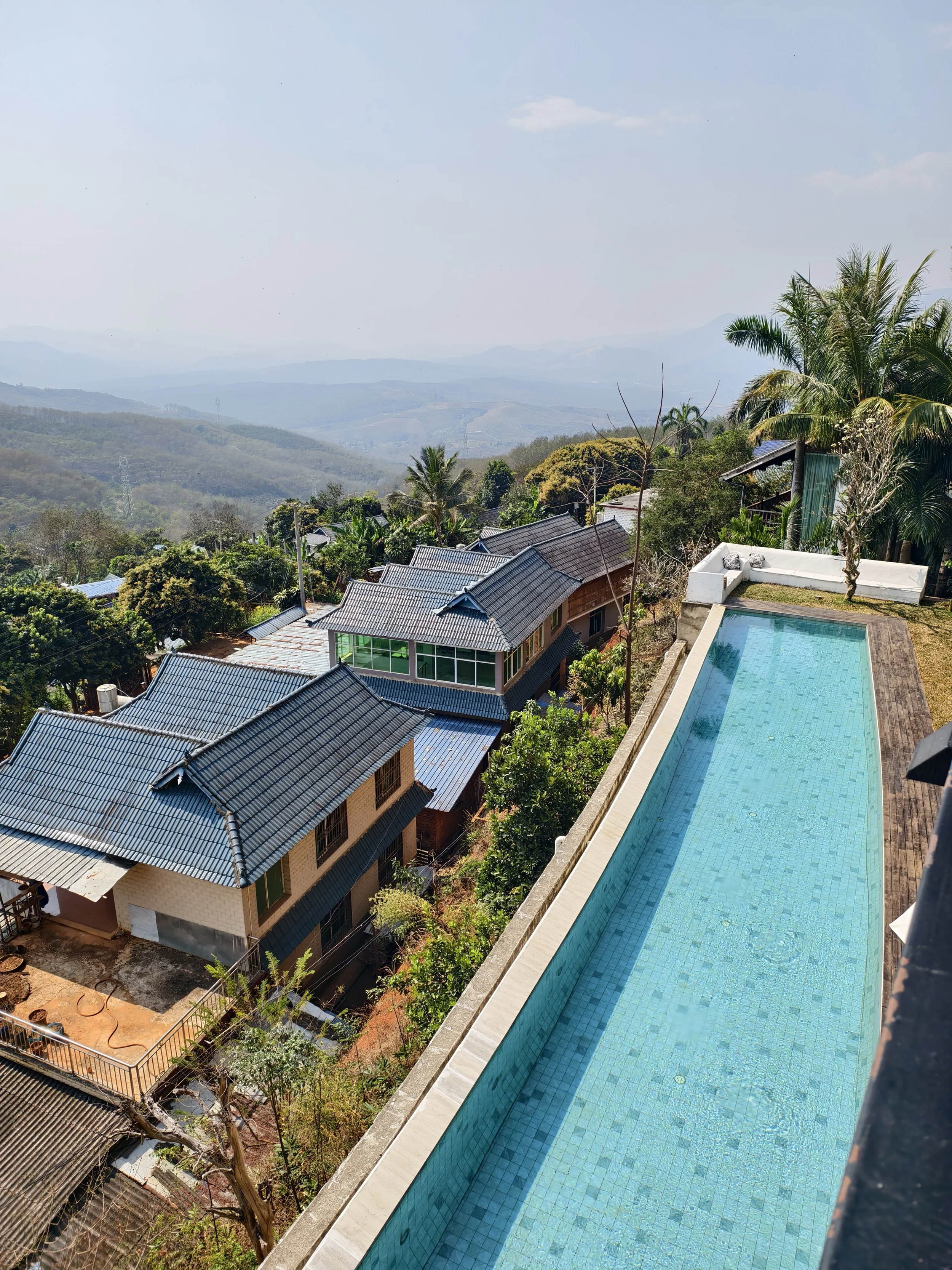 A long swimming pool on a hillside with traditional houses and lush greenery, overlooking a mountain landscape in the distance.