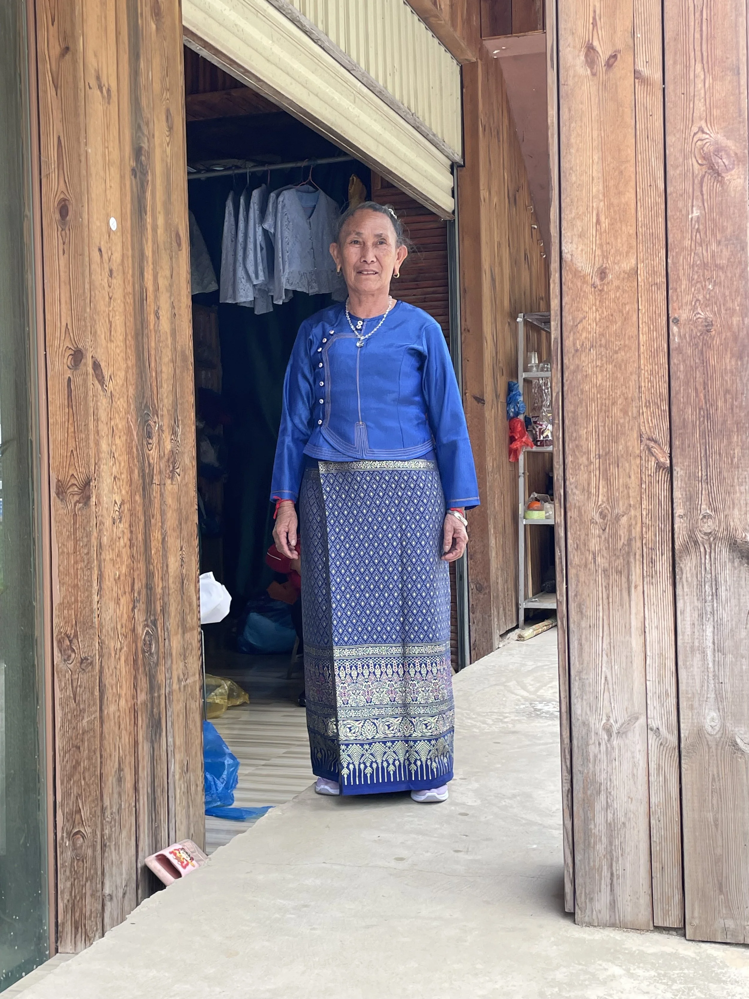 An elderly woman standing in a doorway, wearing traditional blue and patterned skirt, with laundry hanging inside the room behind her.
