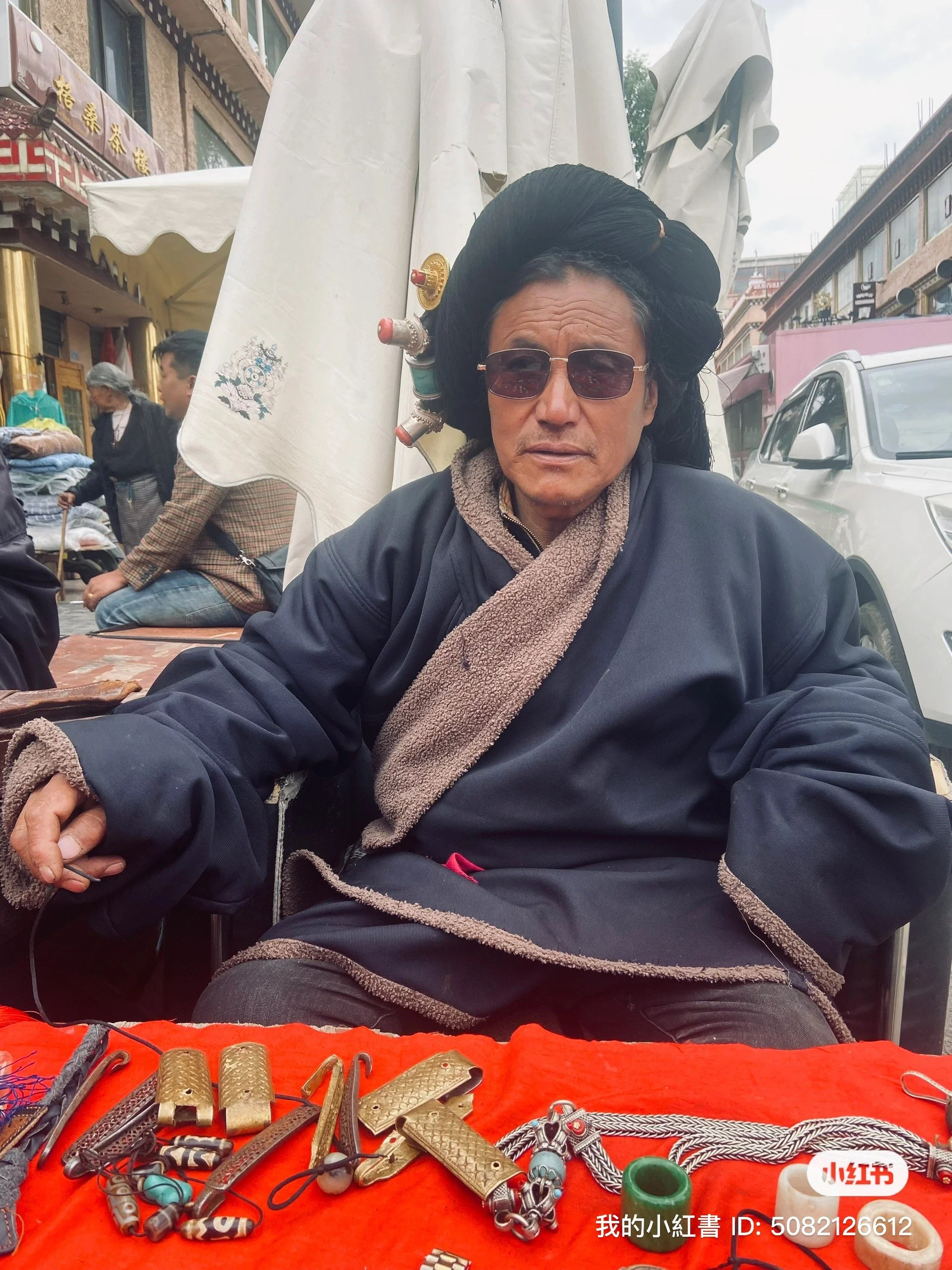 A man with long black hair styled with hair rollers, wearing sunglasses and a black jacket. He is sitting at a market stall displaying various metal jewelry and accessories on a red cloth. Background shows other market vendors, people, and street sce