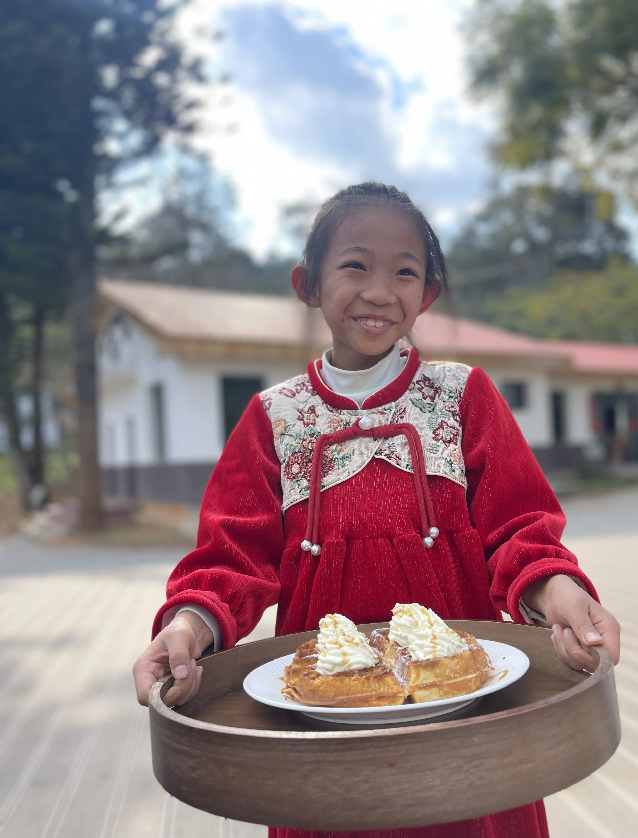 A young girl in a red dress holding a tray with a plate of dessert, smiling outdoors with trees and a house in the background.