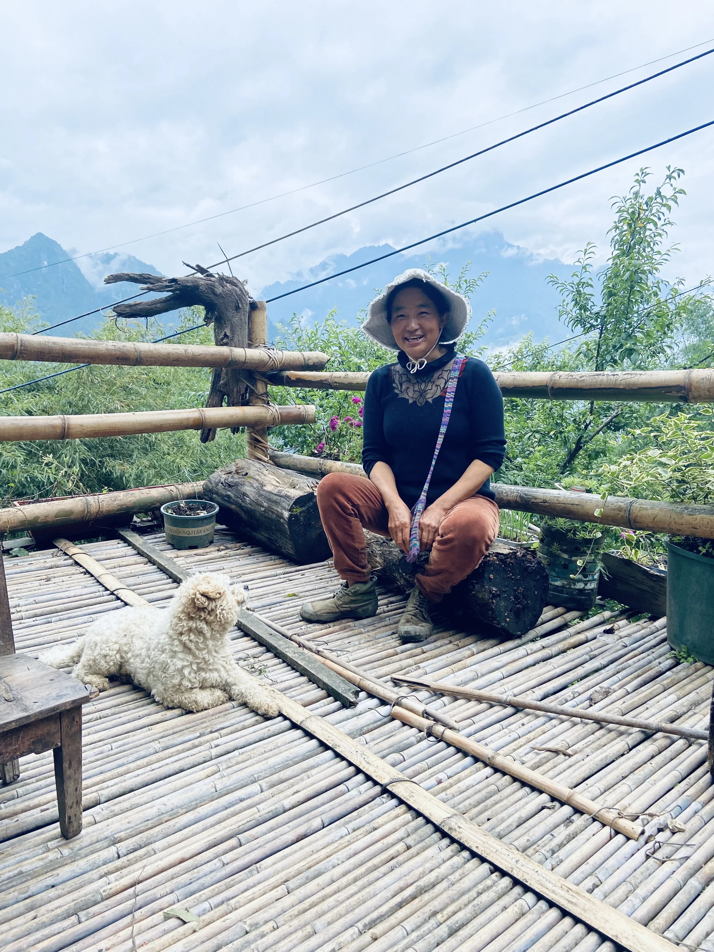 A woman wearing a hat and sitting on a bamboo platform outdoors with a white poodle dog lying next to her, surrounded by mountains and greenery.