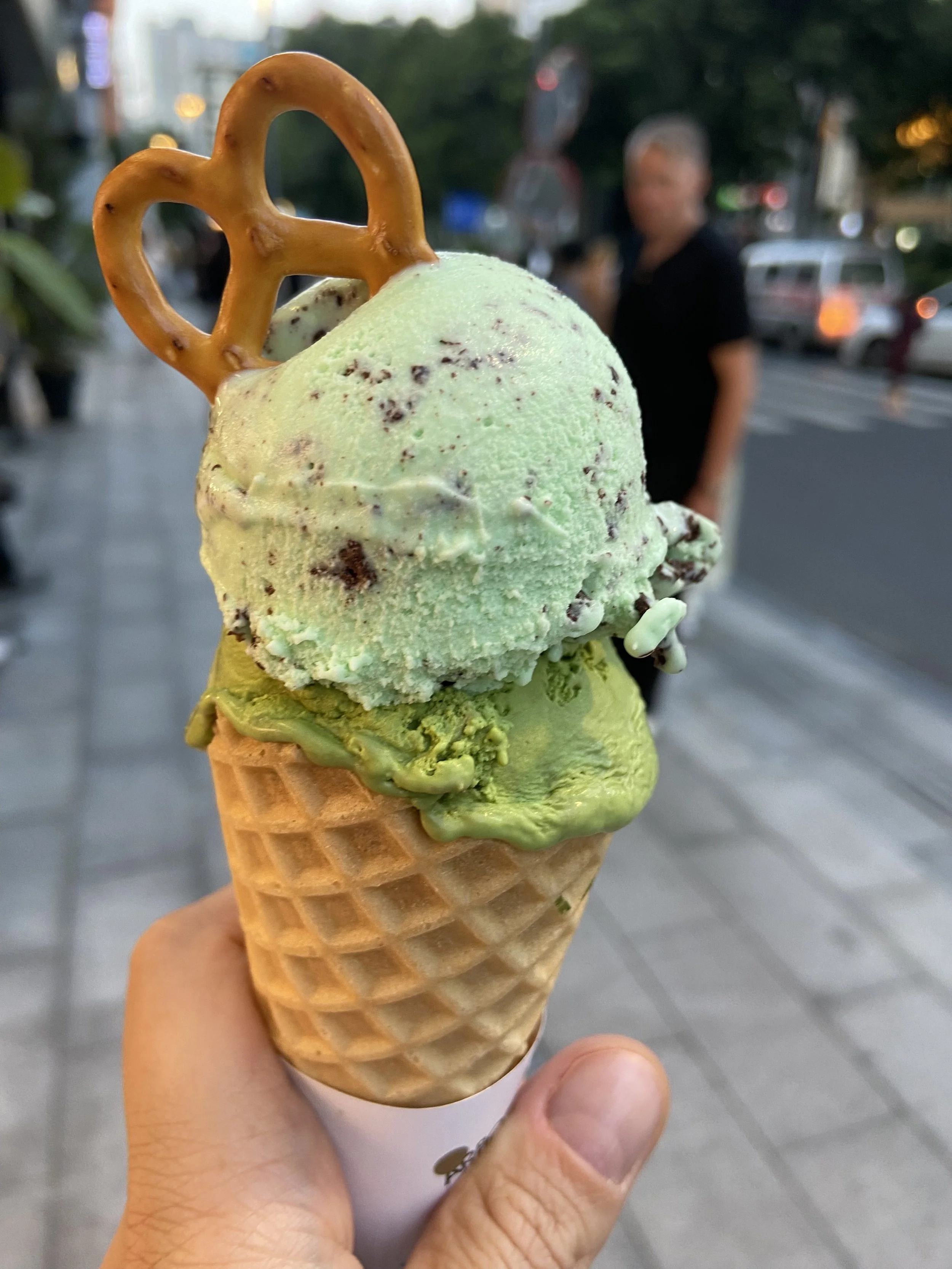 A hand holding a waffle cone with green and white ice cream topped with a pretzel shaped cookie, outdoors on a city sidewalk.