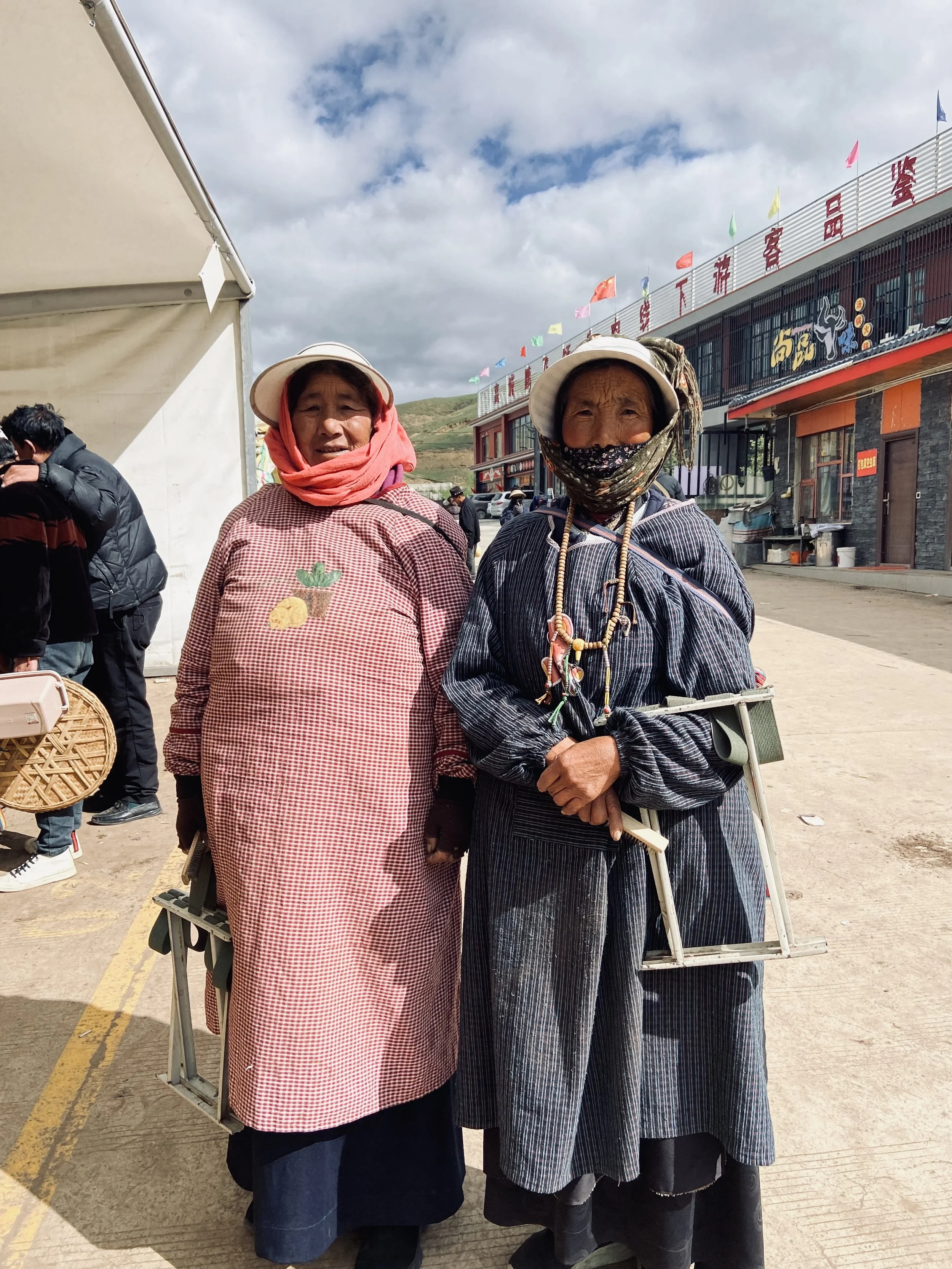 Two elderly women dressed in traditional clothing standing outdoors, one wearing a pink checkered garment with a cartoon character patch, the other in a dark striped outfit, both with hats and masks, holding folding ladders.