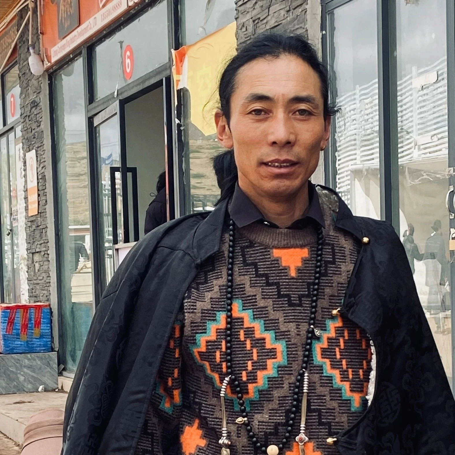 A man standing outside a building with large glass windows, wearing a patterned sweater, a black jacket, and necklaces, with a smile and dark hair tied back.