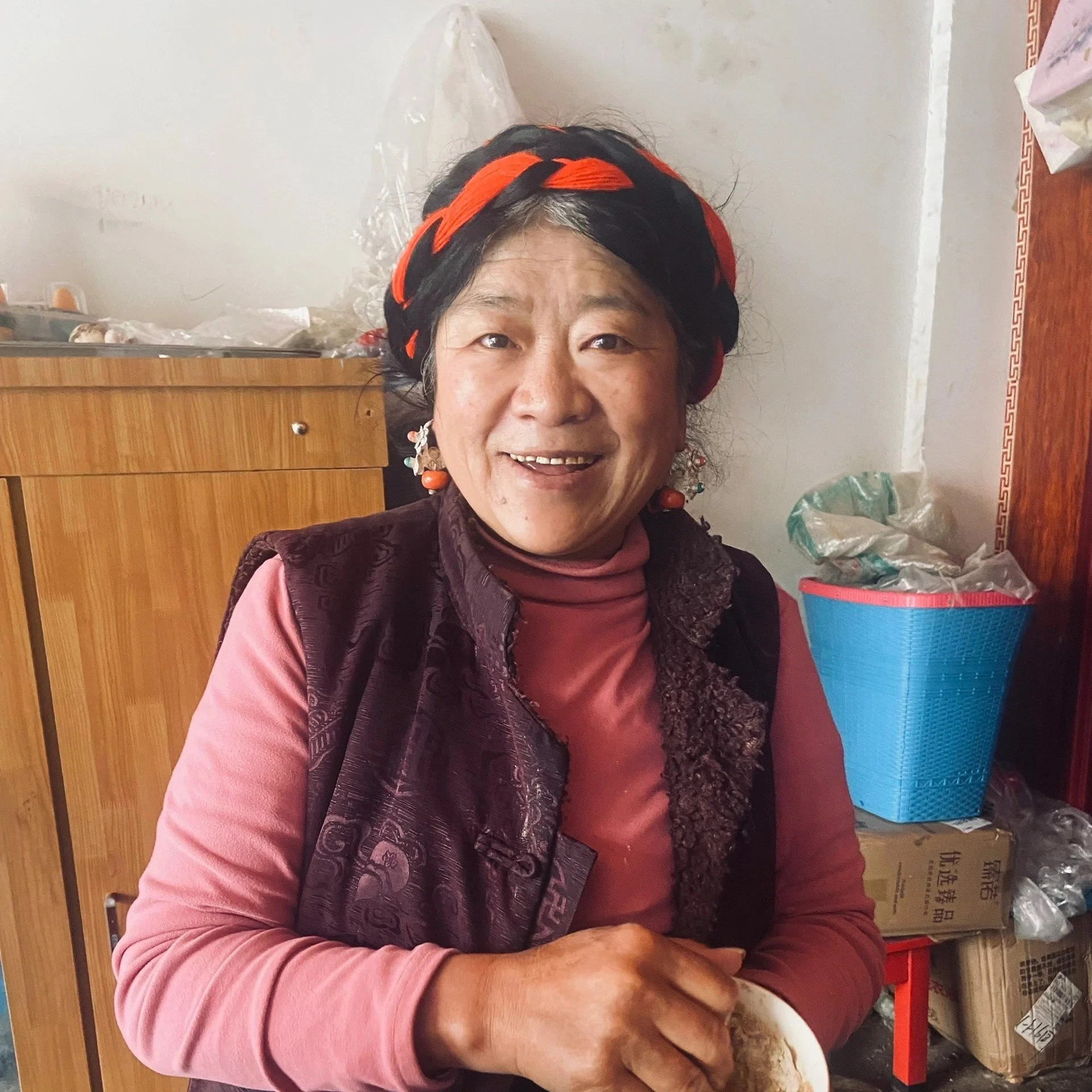 Smiling woman wearing a black and red headband, earrings, a pink turtleneck, and a dark vest, sitting indoors with a wooden cabinet and boxes in the background.
