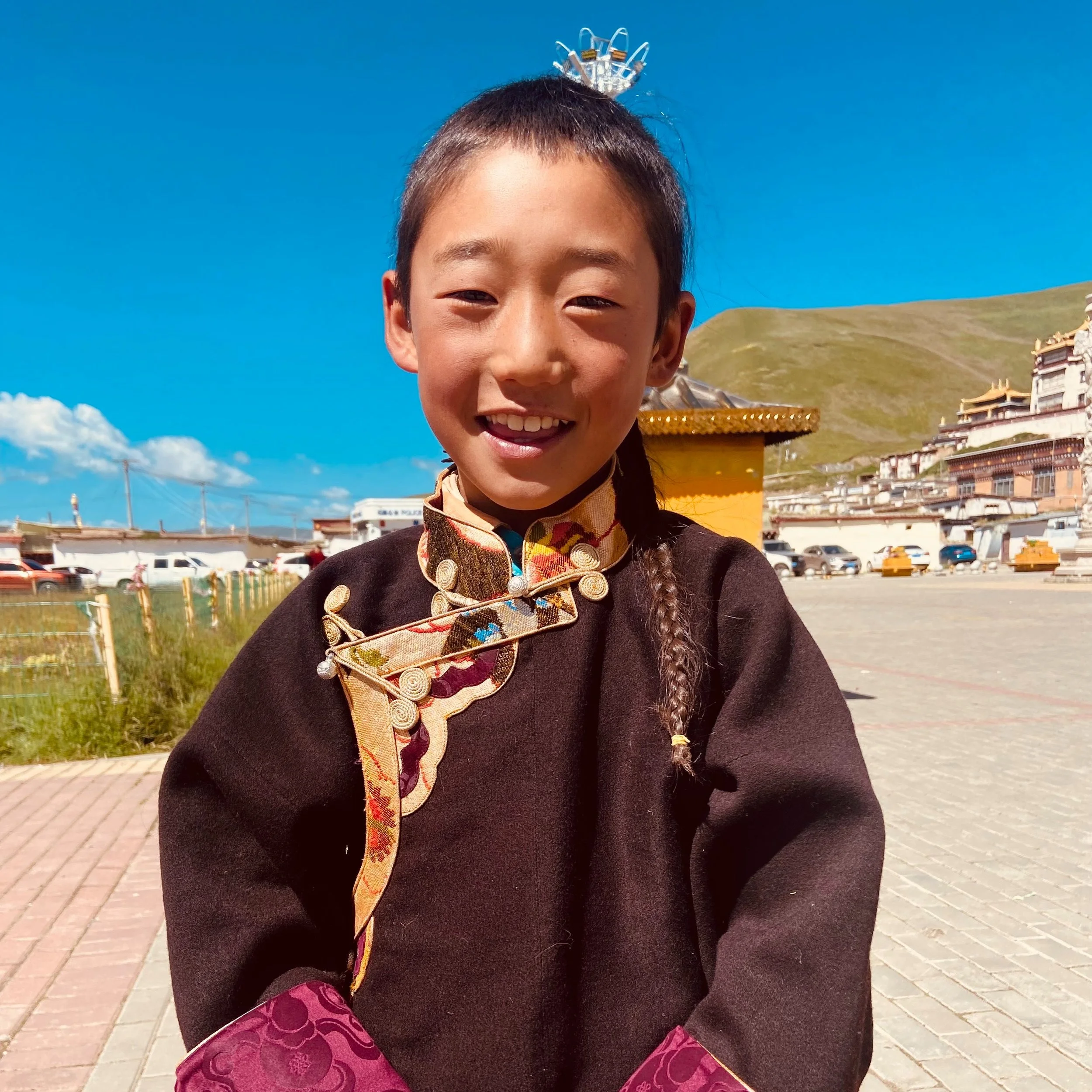 Smiling young girl in traditional clothing standing outdoors with a Buddhist monastery and hillside in the background.