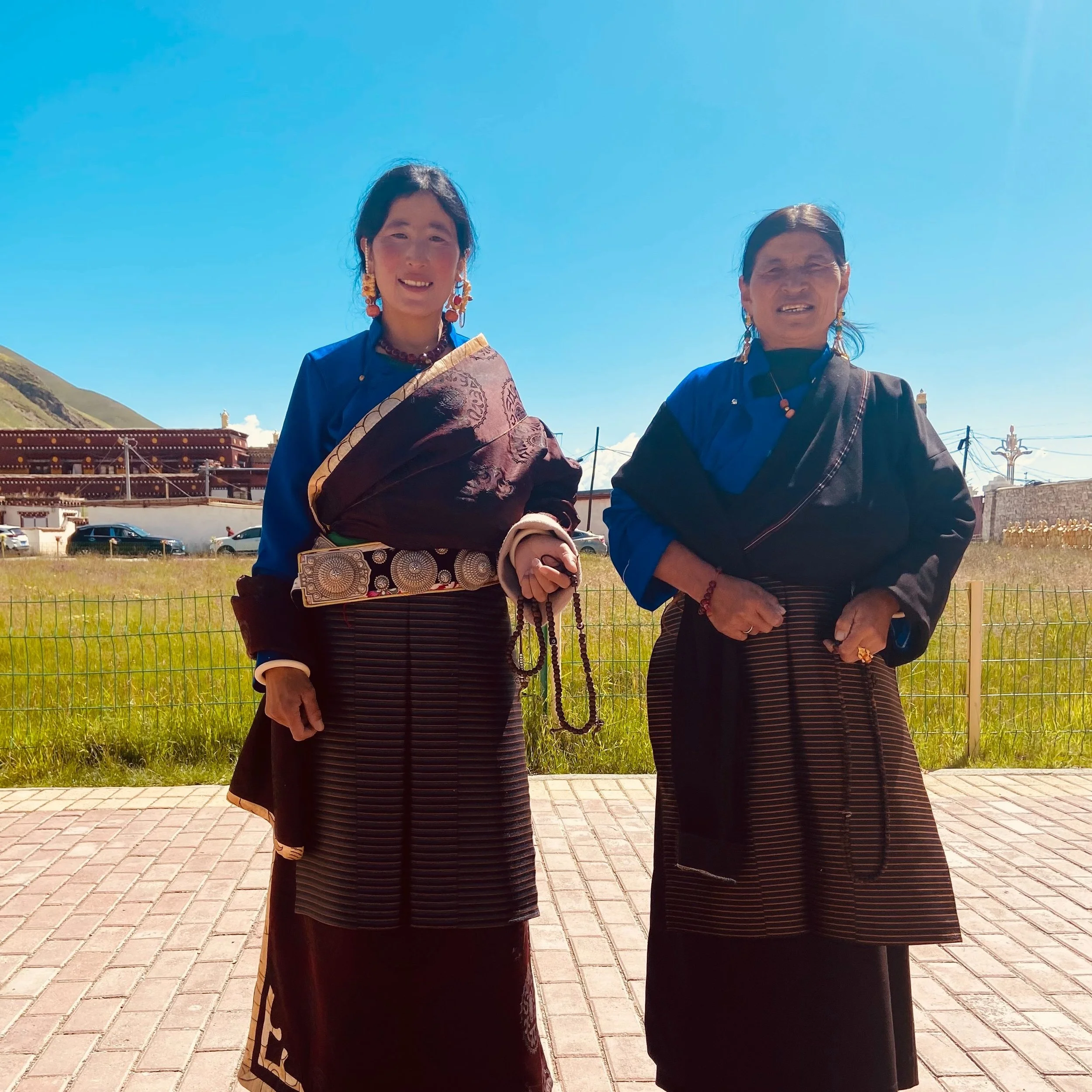 Two women in traditional Tibetan attire standing outdoors on a sunny day with a grassy field and a building in the background.