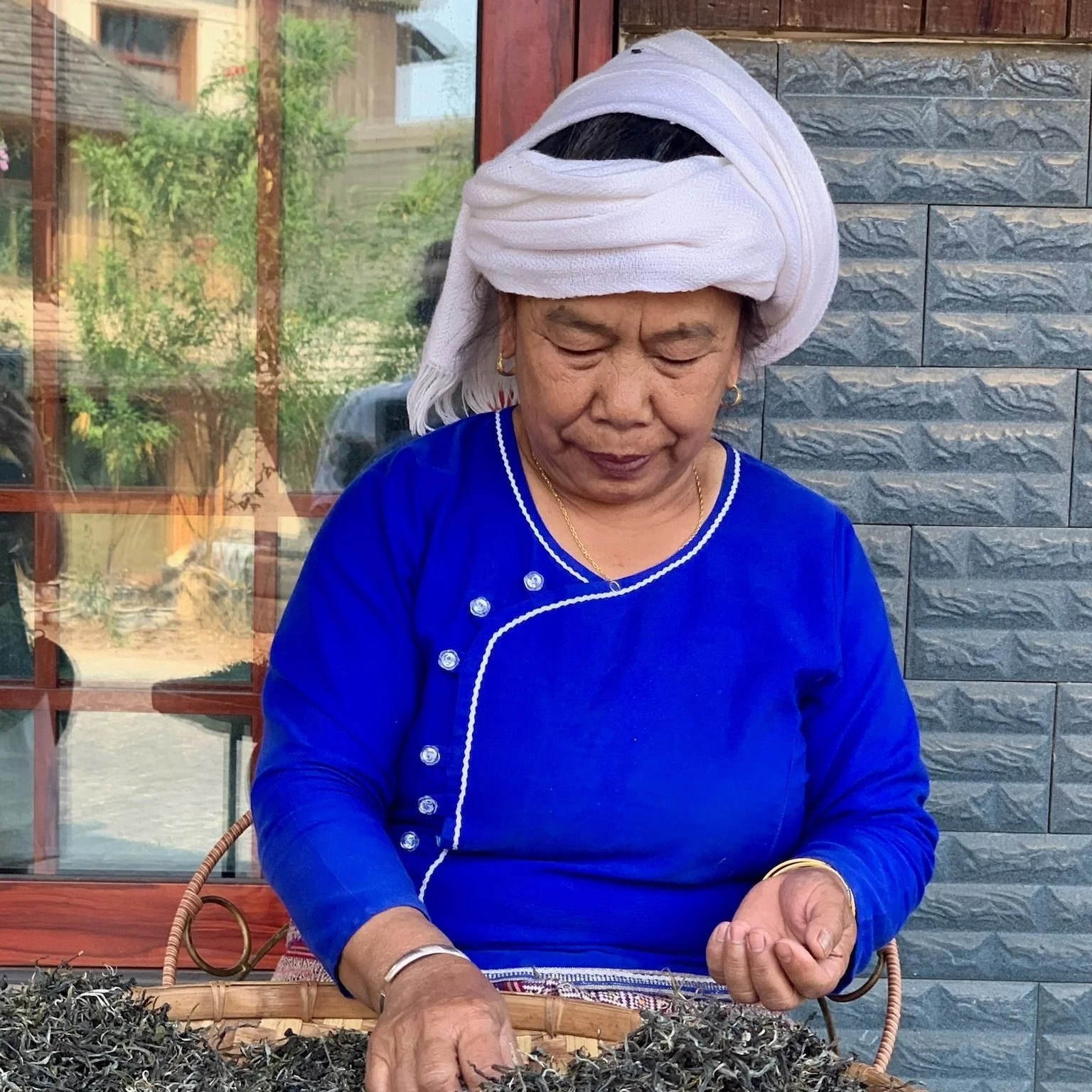 An elderly woman dressed in a bright blue traditional garment with white embroidery and a white headscarf, working with dried herbs or tea leaves in a basket outdoors near a building with a stone wall and window.