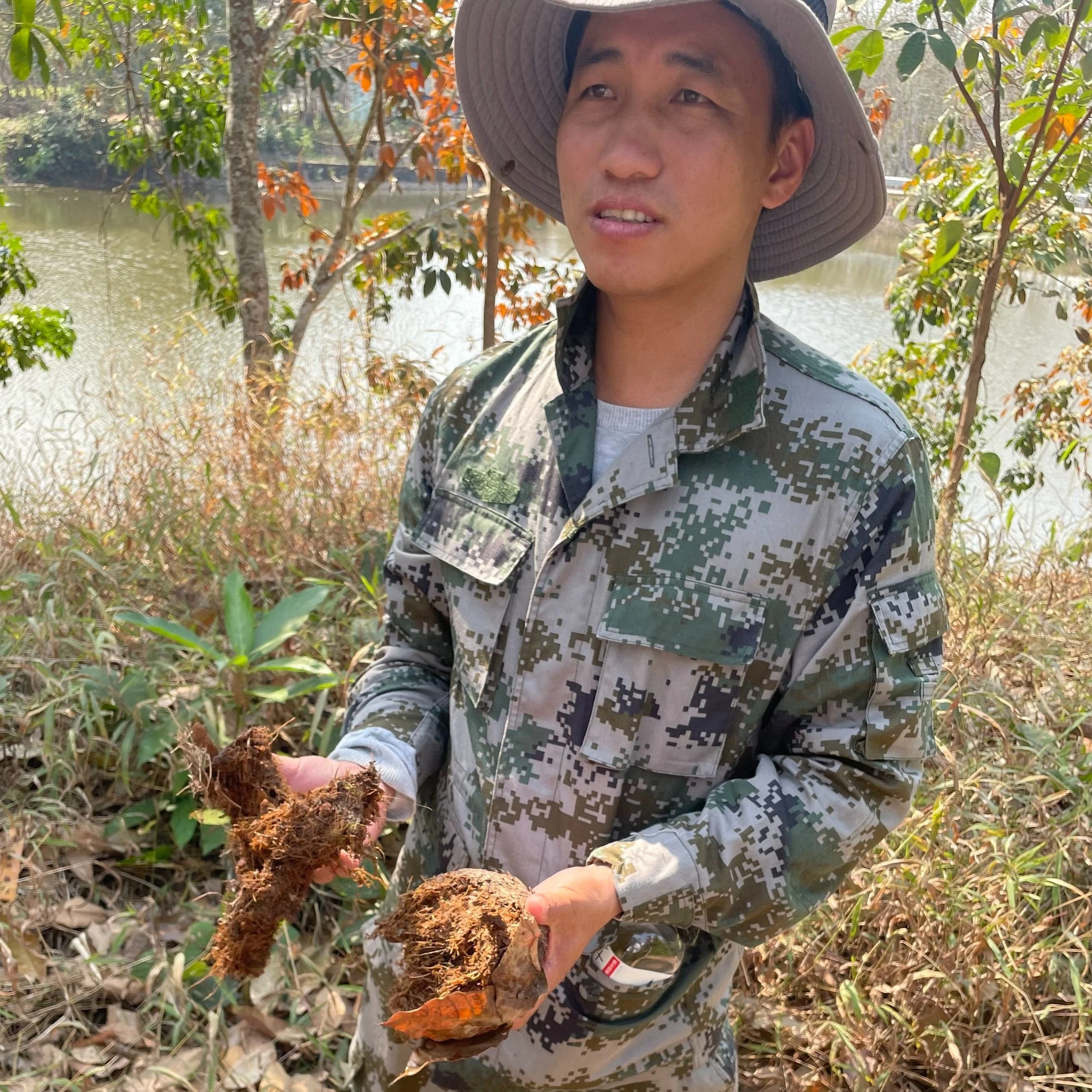 A man in camouflage military uniform holding roots and a large orange fungus or plant part outdoors near a river, with trees and dry leaves on the ground.