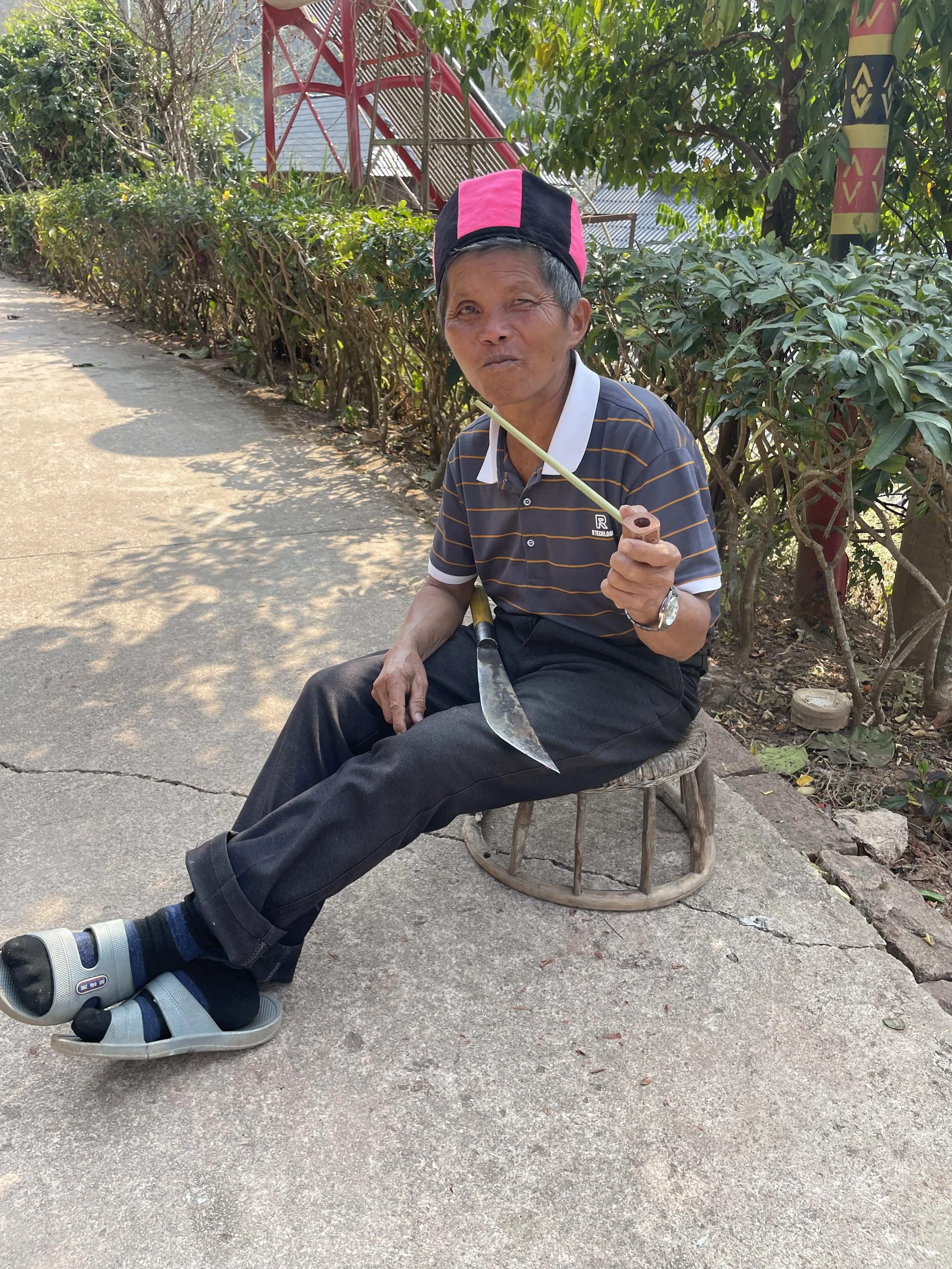 An elderly man sitting on a small wooden stool holding a knife and a stick with a humorous expression. He's wearing a striped polo shirt, dark pants, sandals, and a black and pink hat. The background shows greenery and an outdoor setting with sunligh