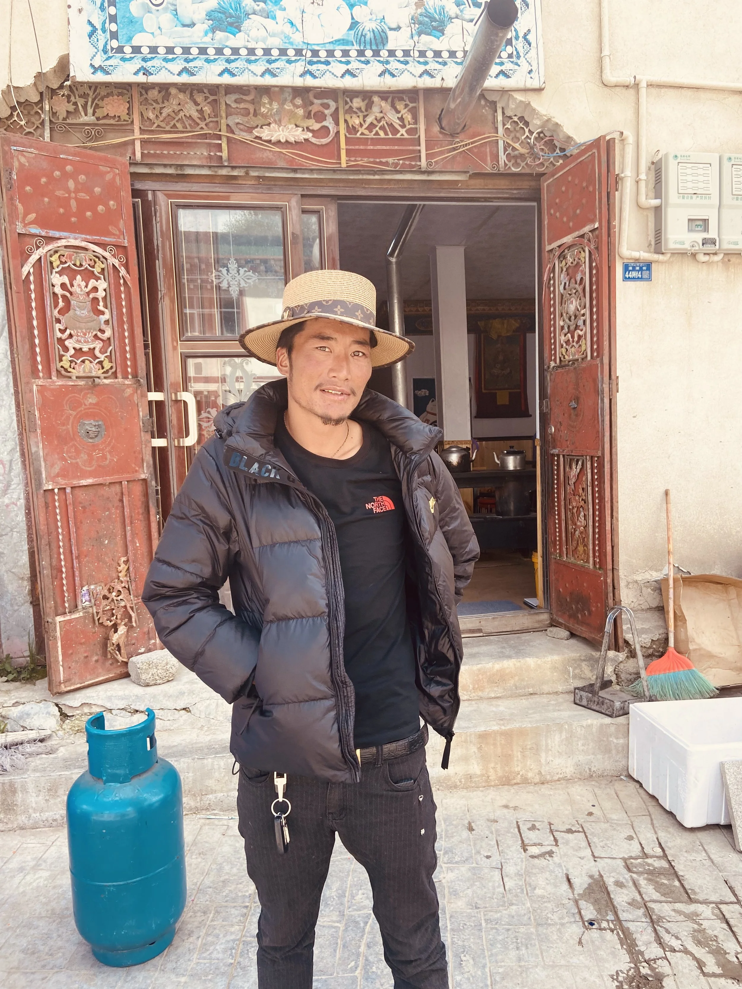 Man standing outside a building with open iron gates, wearing a black jacket, black t-shirt, black pants, and a straw hat, with a blue propane tank nearby.