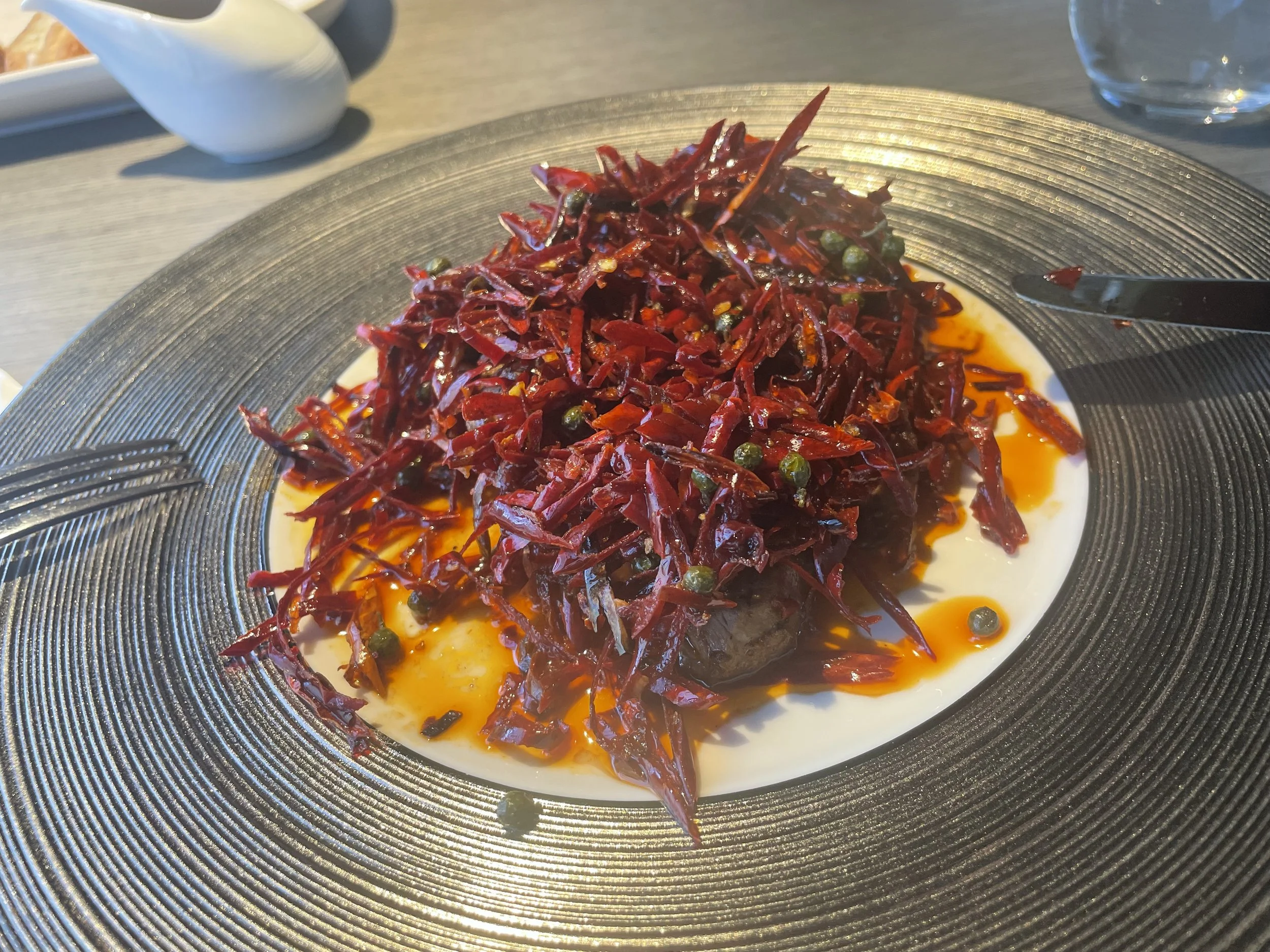 A black plate with a salad of shredded red vegetables, possibly beets, topped with small green peppercorns and drizzled with orange sauce, set on a light-colored table.