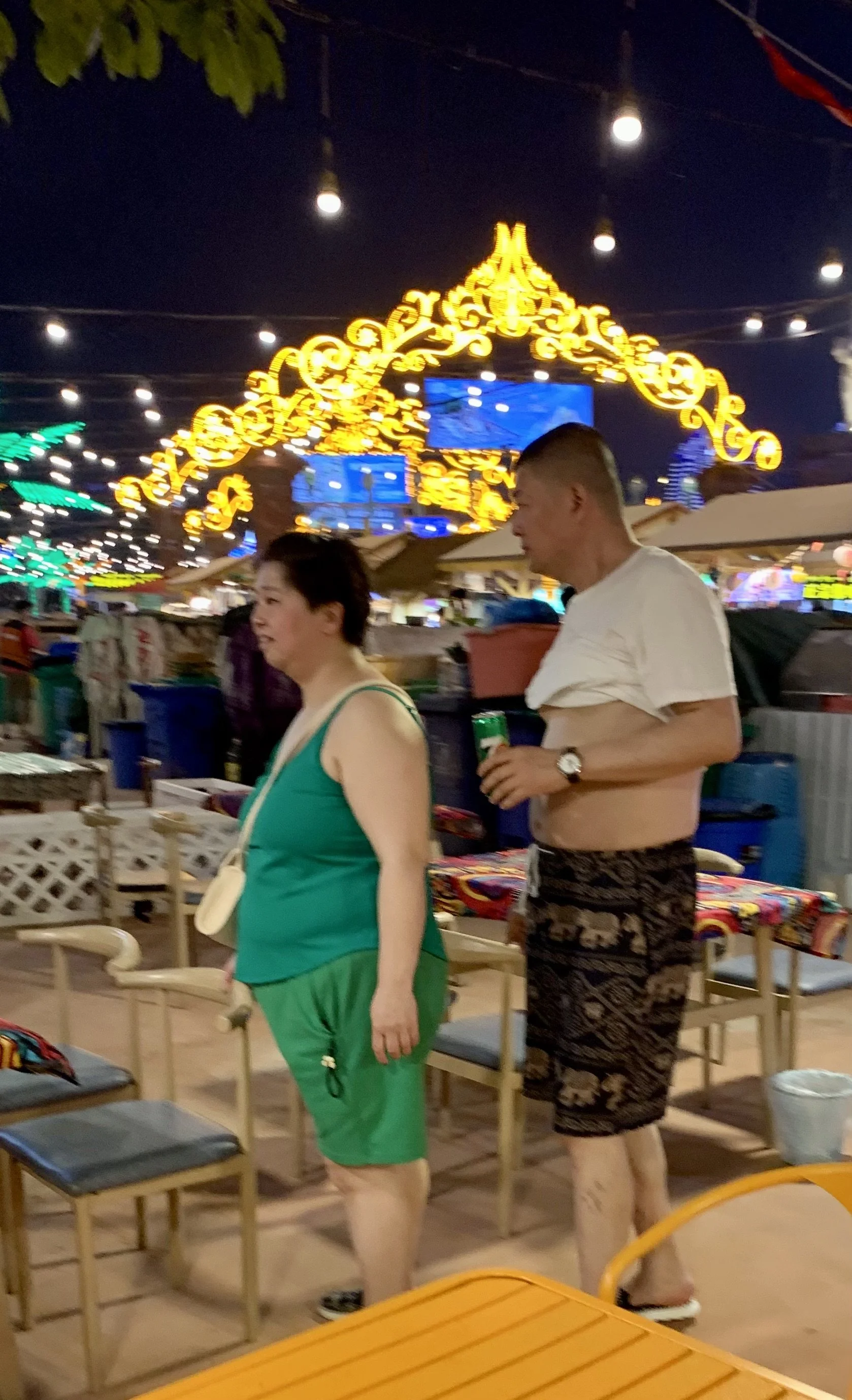 Two people in casual summer clothing standing at an outdoor dining area at night, with bright decorative lights and colorful booths in the background.