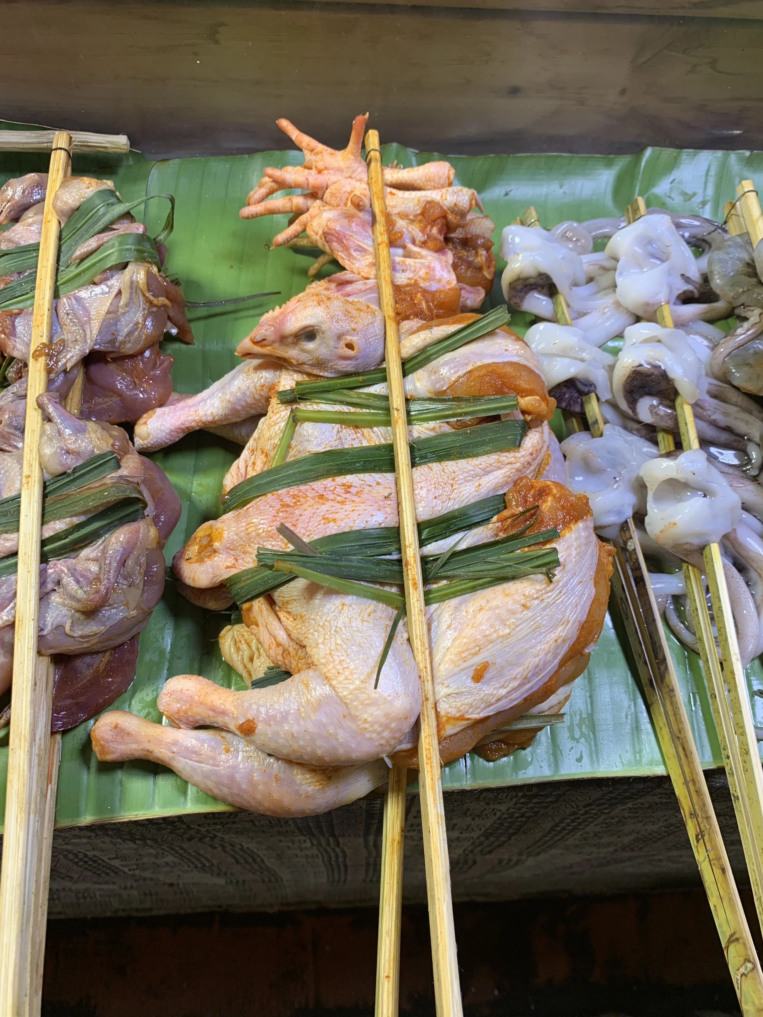 Skewers of raw chicken, frog legs, squid, and chicken hearts arranged on a banana leaf at a market or food stall.