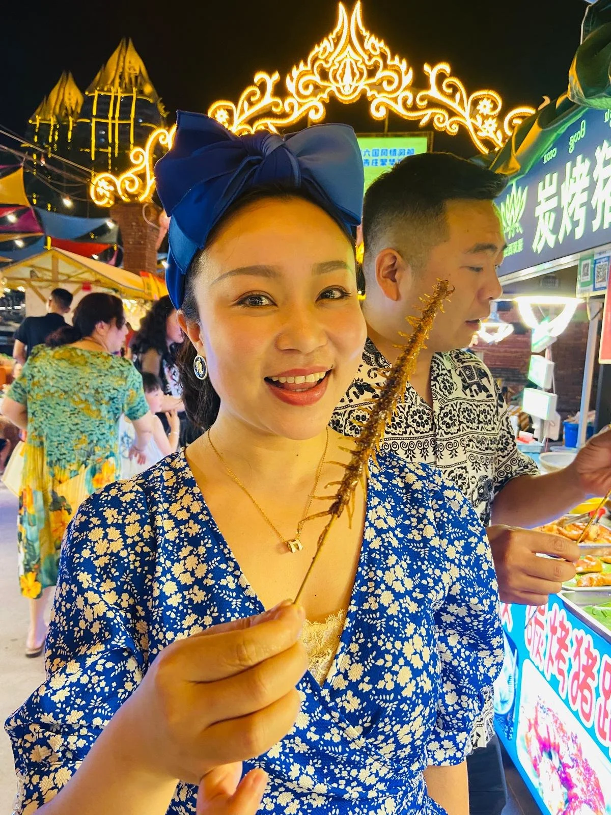 A woman smiling at a night market holding a grilled skewer, with bright lights and other people in the background.