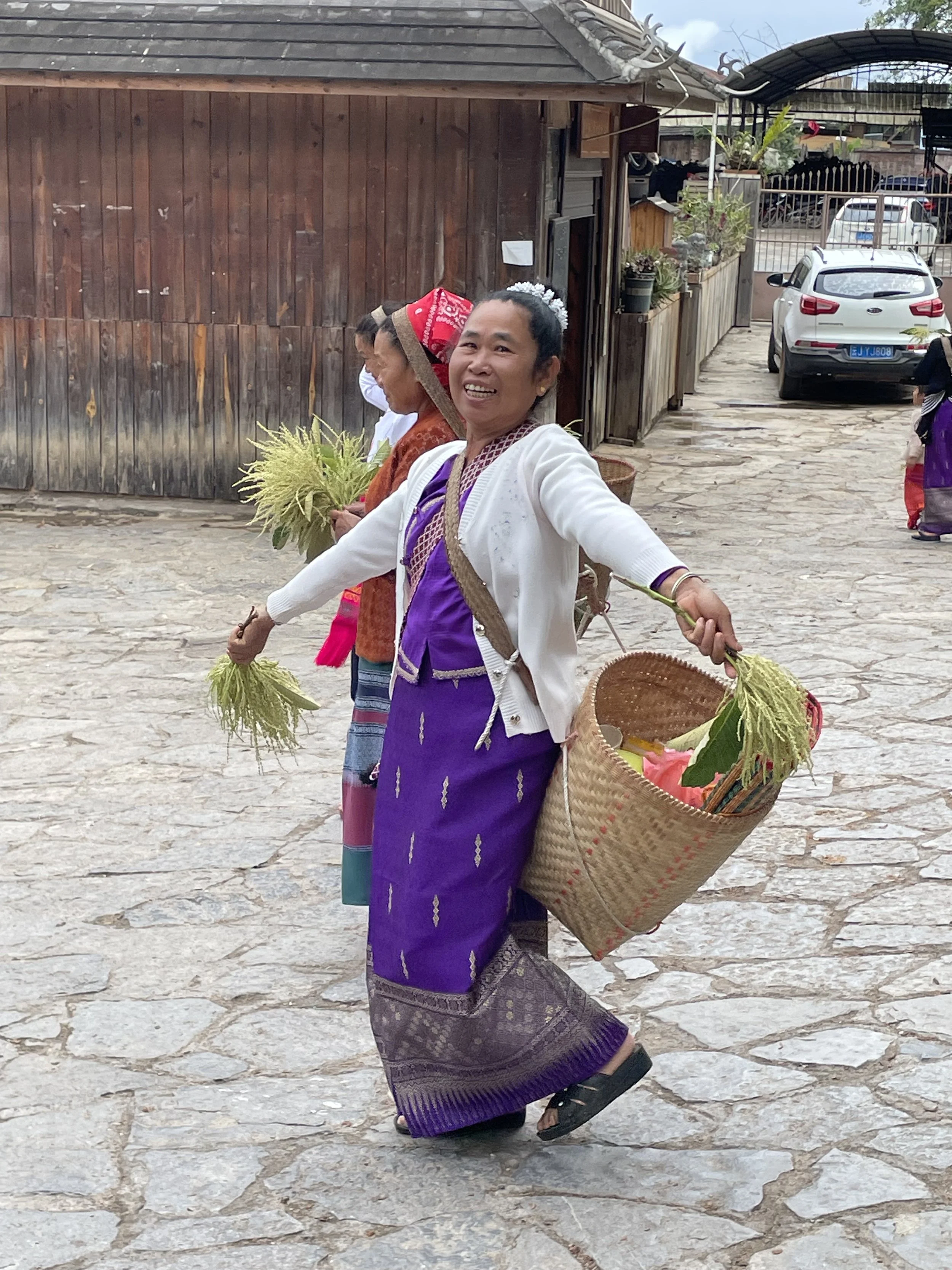 Women in traditional clothing smiling and holding plants and a woven basket, walking on a cobblestone street, with a wooden building and cars in the background.