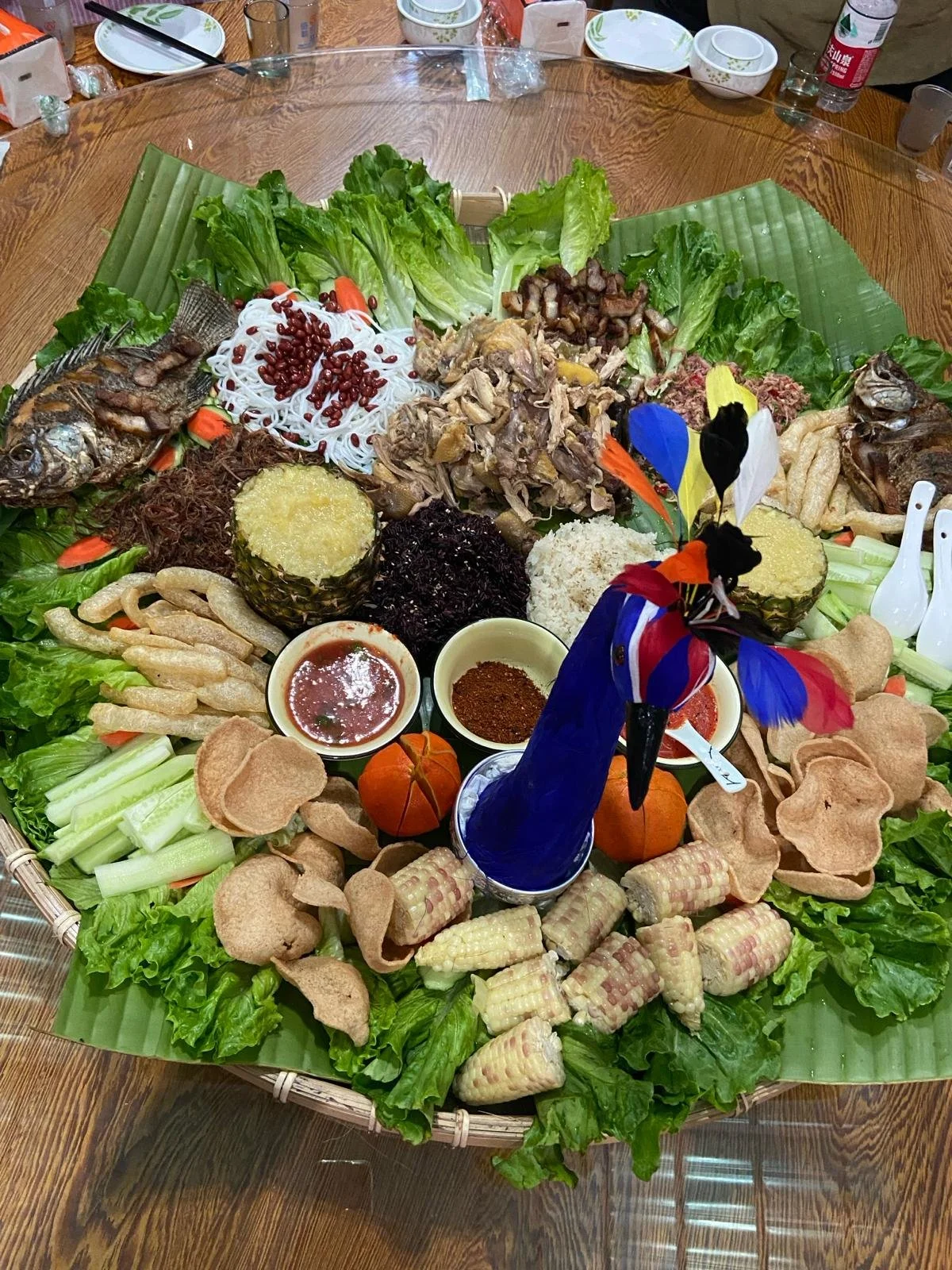 A large, colorful Thai-style food platter on a round table with various vegetables, grilled fish, rice, and sauces arranged around a decorative rooster-shaped centerpiece.
