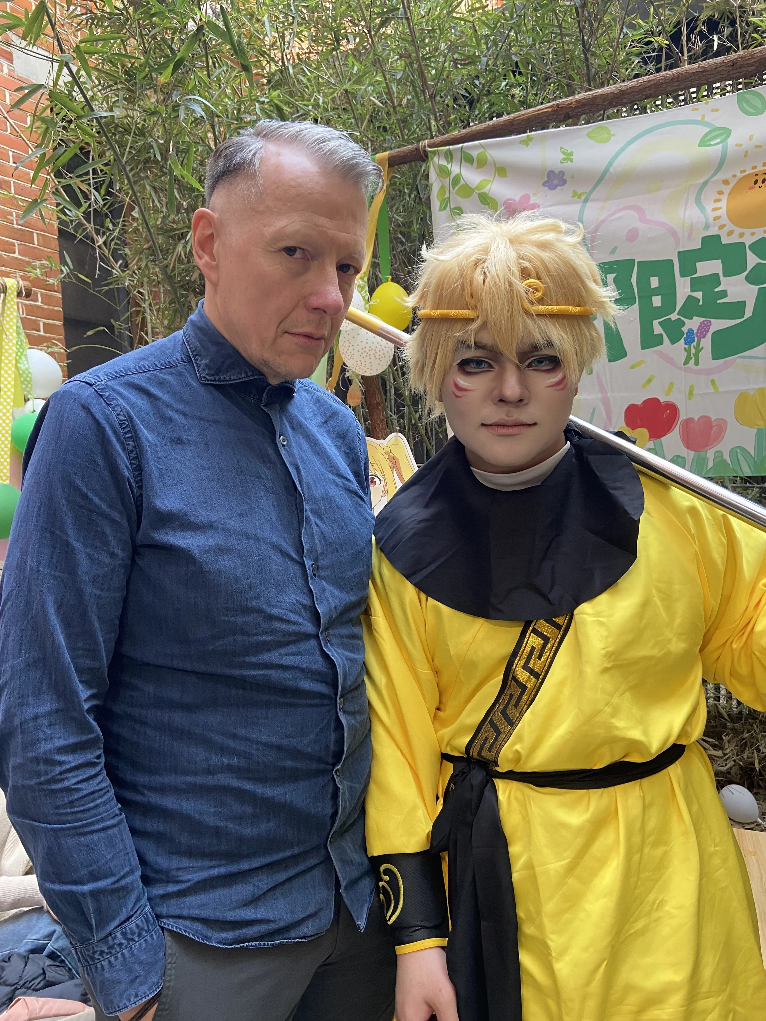 Two men standing together, one dressed casually in a blue denim shirt, and the other in a bright yellow costume with painted face, yellow hair, and a staff, at an outdoor event decorated with colorful balloons and a banner.