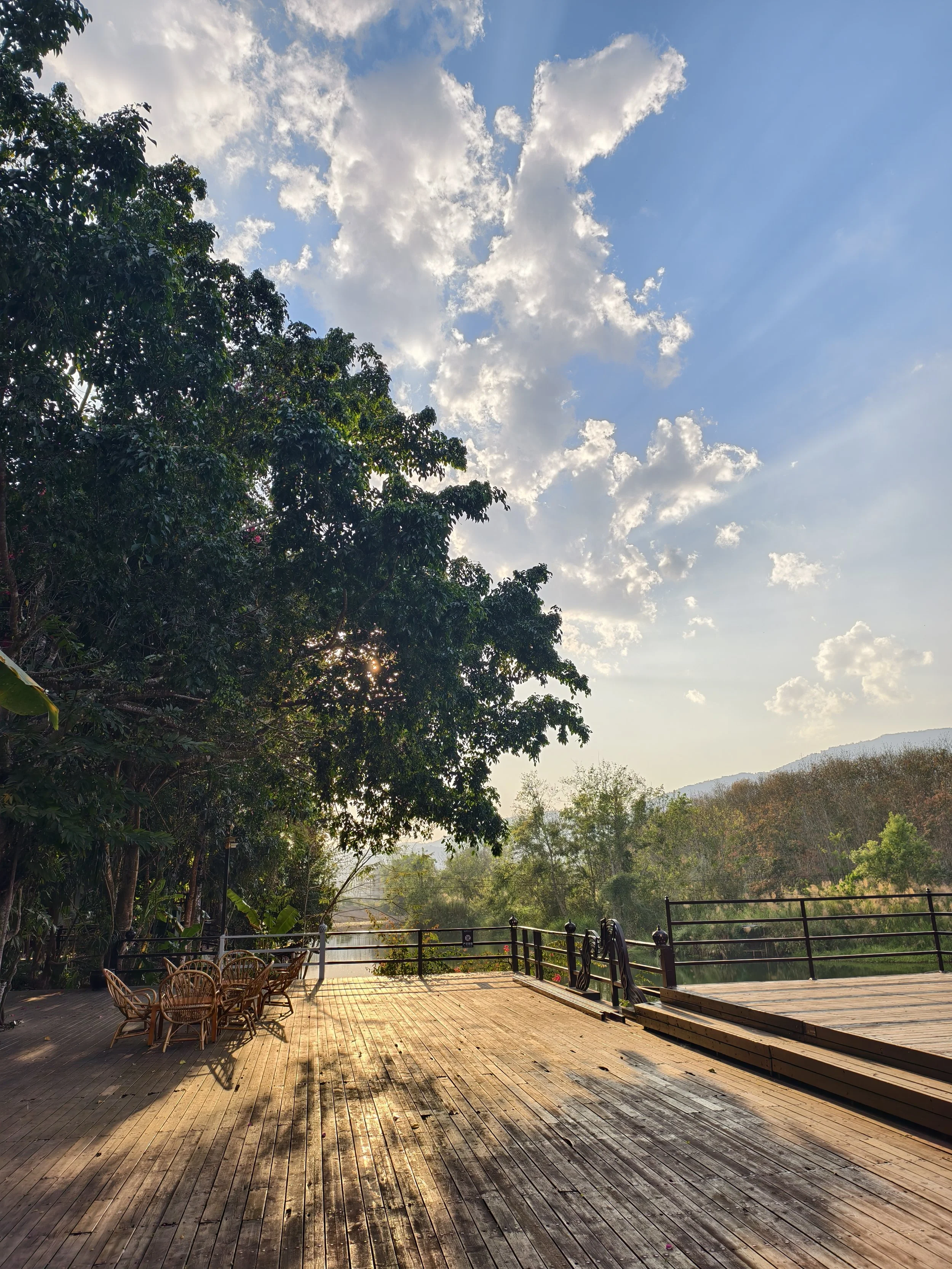 Wooden deck overlooking a river with green trees and mountain in the distance, sky with fluffy clouds, and sunlight casting shadows, outdoor setting.