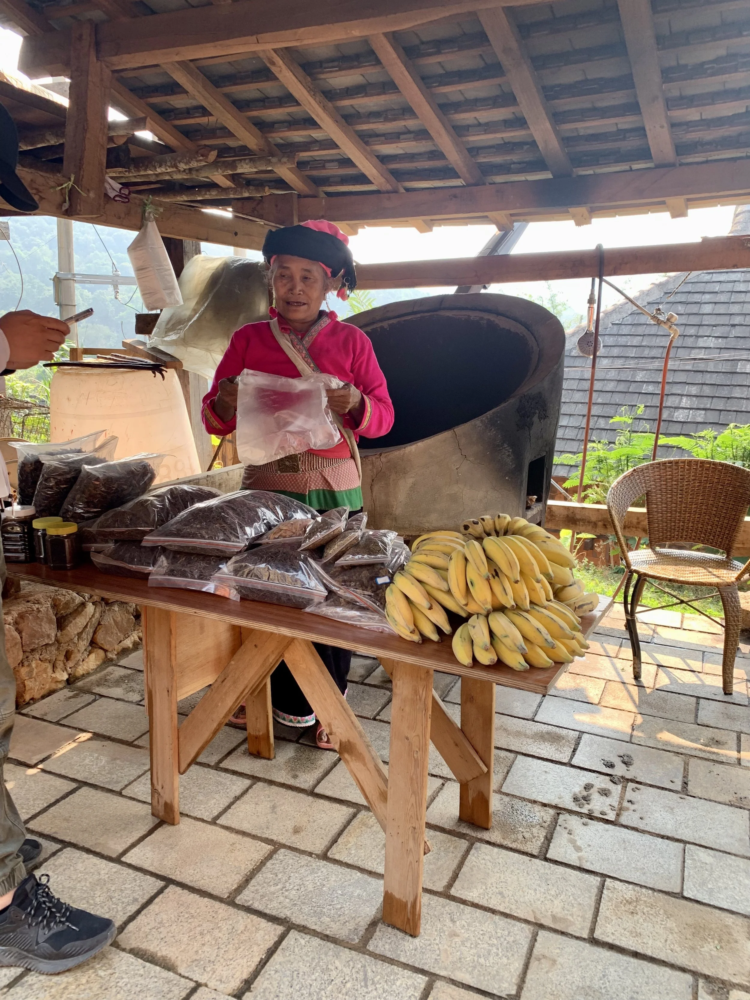 A woman in traditional attire selling bananas and packaged goods at a market stall with a brick floor, wooden roof, and a large black oven in the background.