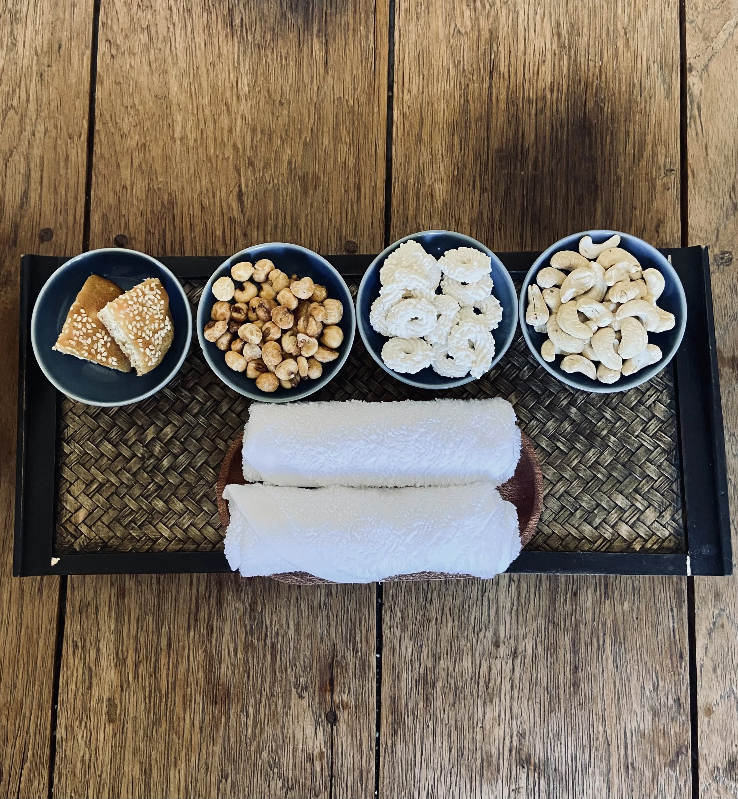 A black tray with four small bowls containing snacks, two rolled white towels, on a wooden table. The snacks include sesame-coated sweets, roasted chickpeas, crispy rice cookies, and cashew nuts.