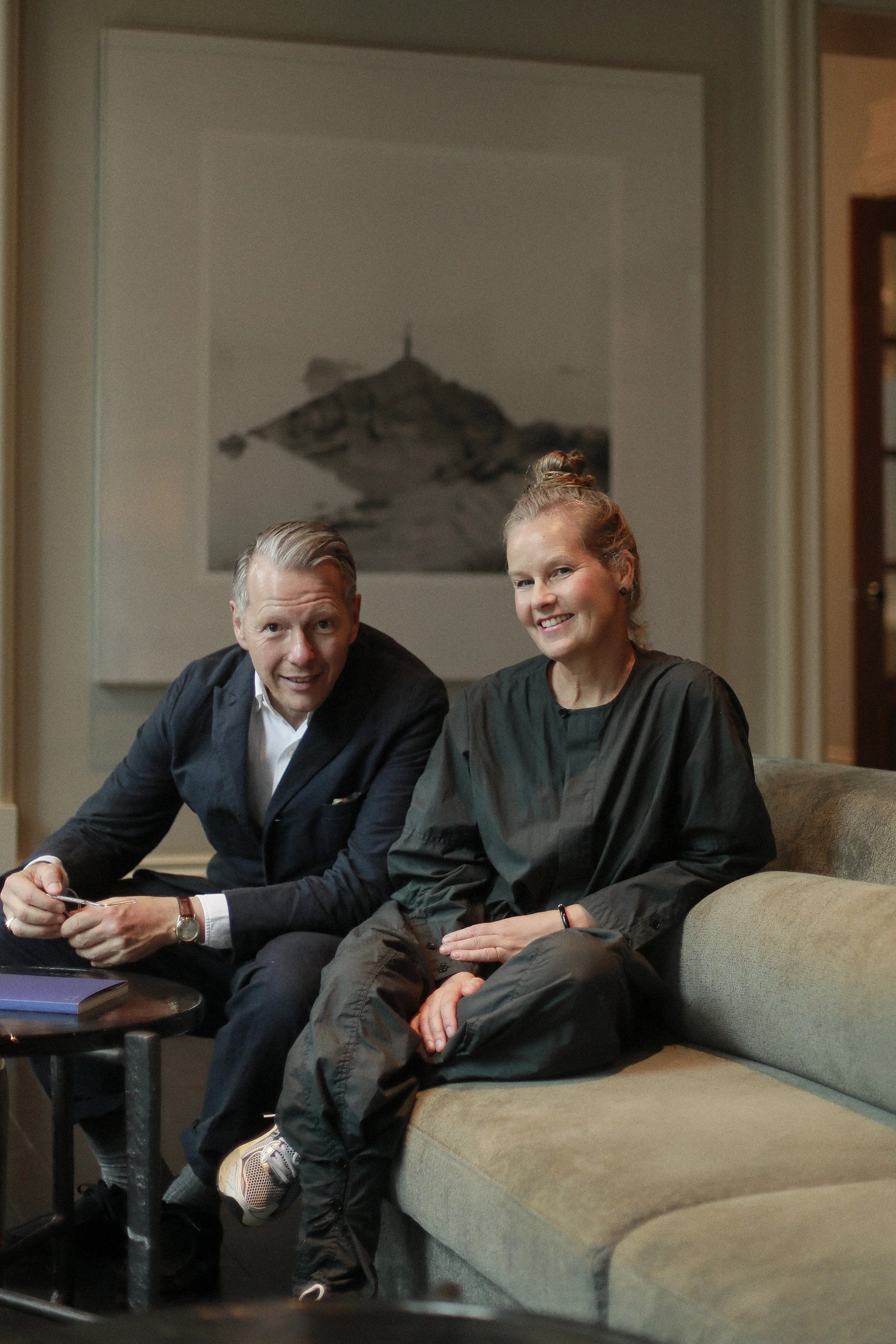 A man and a woman sitting on a couch in a cozy living room, smiling at the camera. The man is wearing a suit and has short, slicked-back hair. The woman is dressed in casual attire with her hair tied back. There is a painting or photograph hanging on the wall behind them, and a small table with books and objects is in front of them.