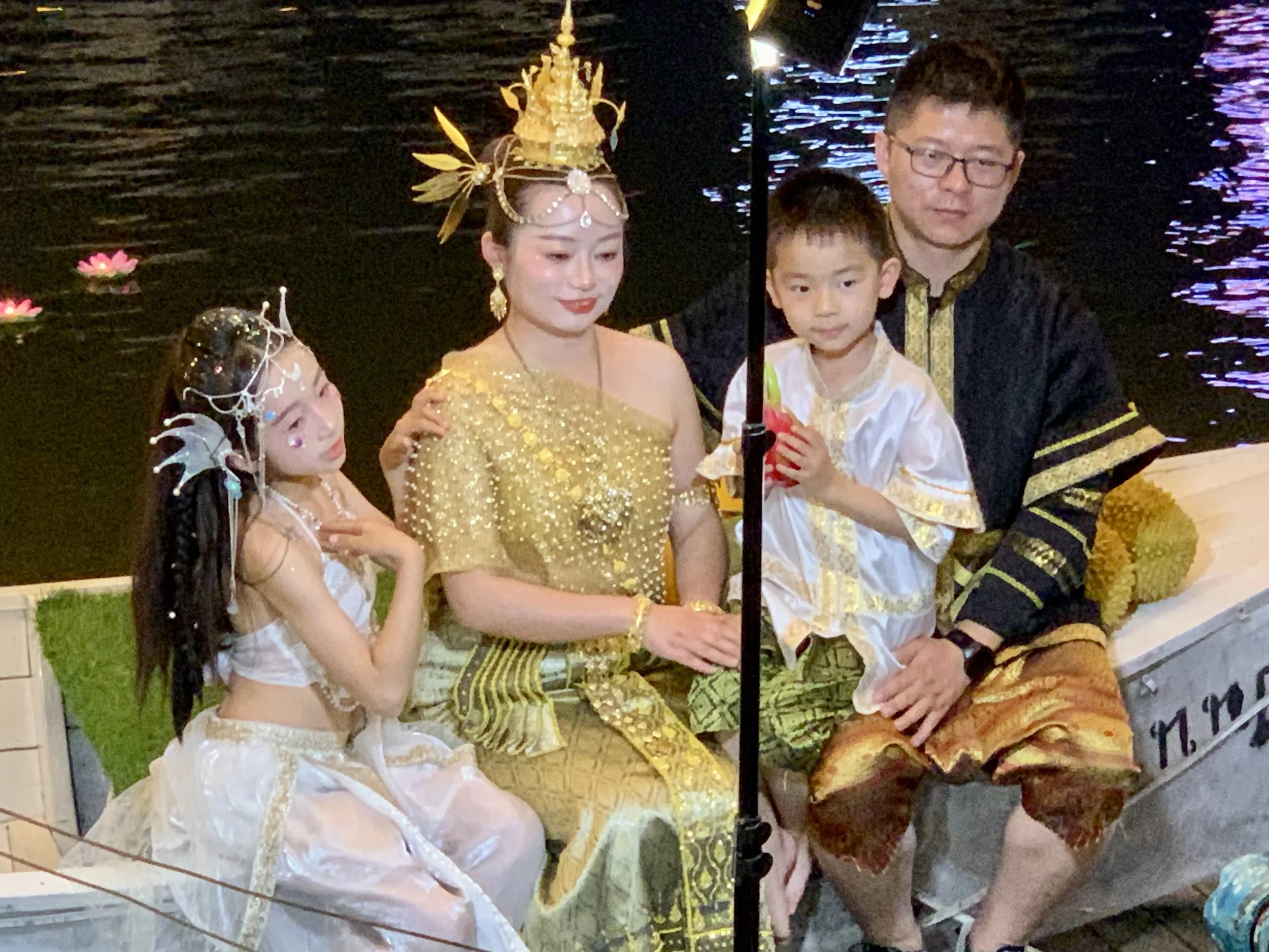 A family dressed in traditional Thai clothing sitting in a boat by a body of water at night, participating in a cultural celebration.