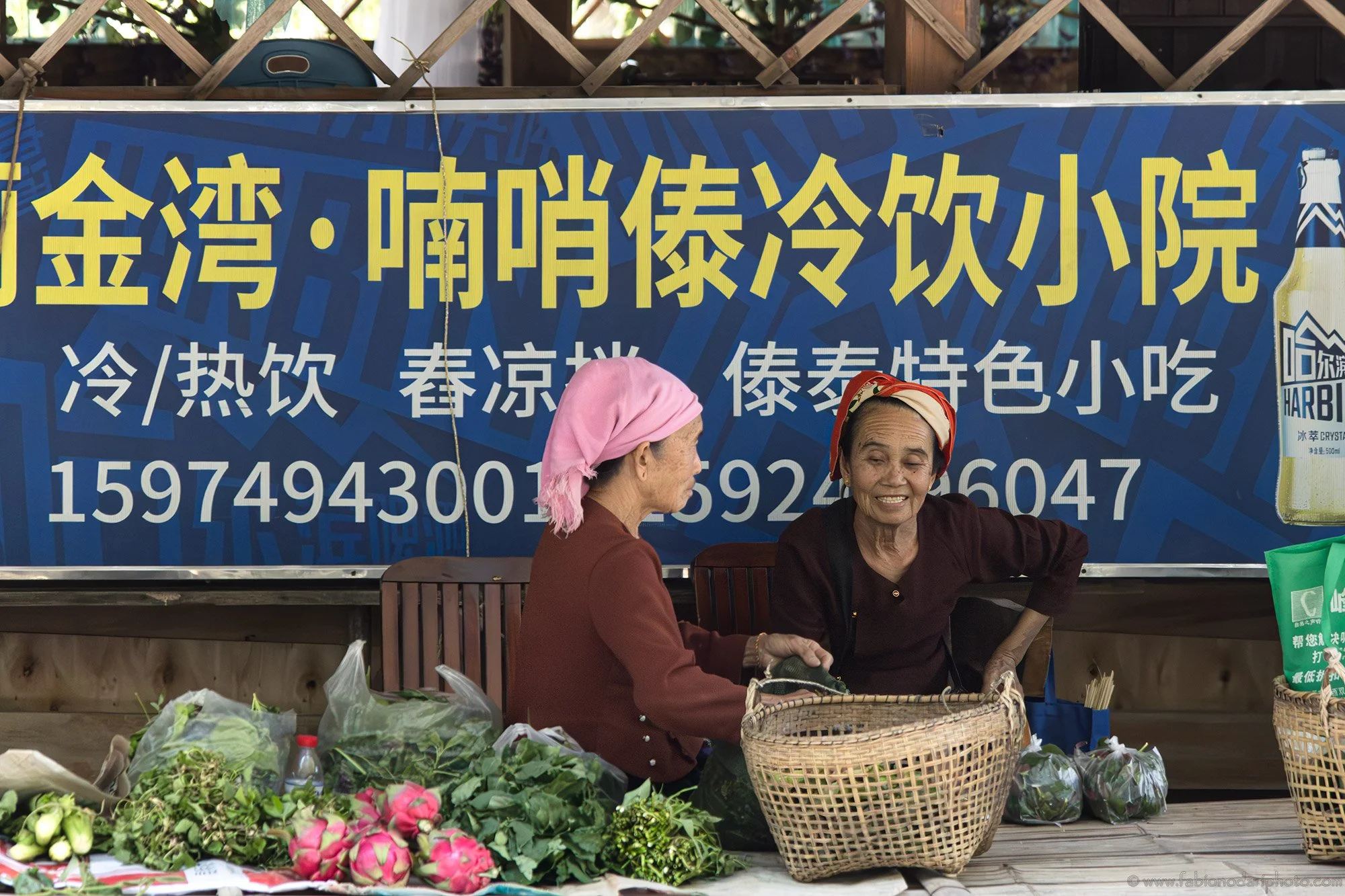 Two women are sitting behind a market stall with vegetables and flowers, talking, with a blue sign with Chinese characters behind them.