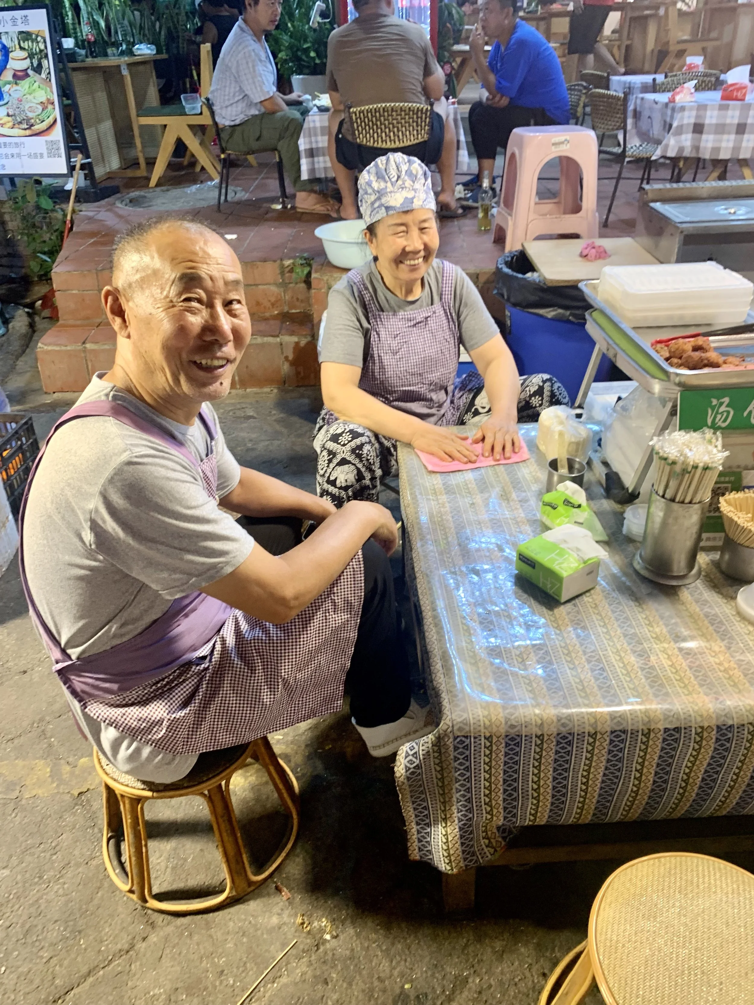Two smiling street vendors, a man and a woman, sitting at a table covered with a patterned cloth, at an outdoor market.