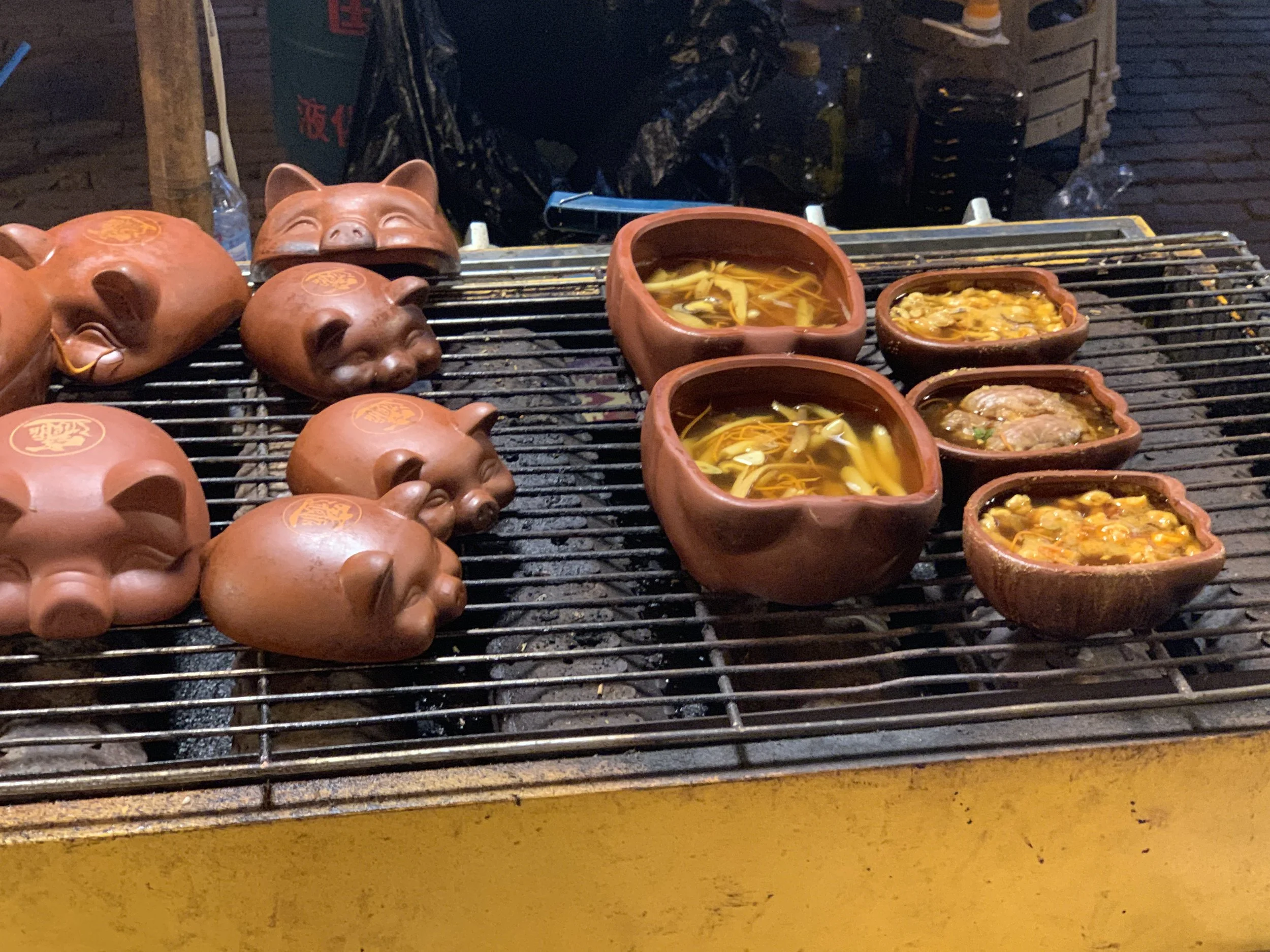 Pig-shaped clay containers and small clay bowls with soup on a grill grate at a street food stall.