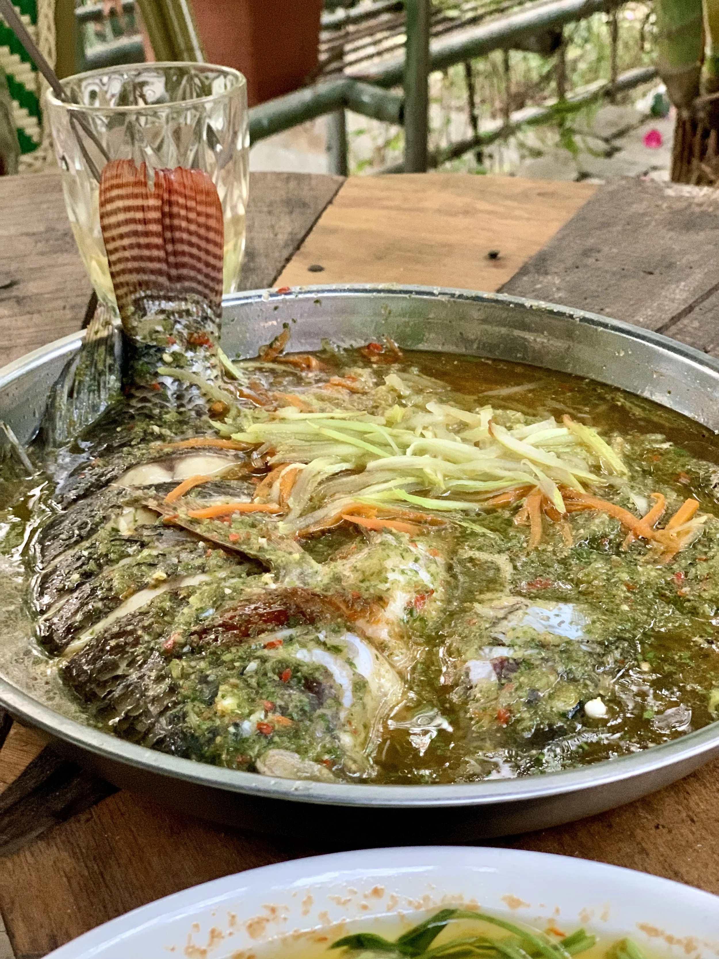 Fresh fish cooked in a green herb sauce on a metal serving dish with shredded vegetables on top, placed on a wooden table.