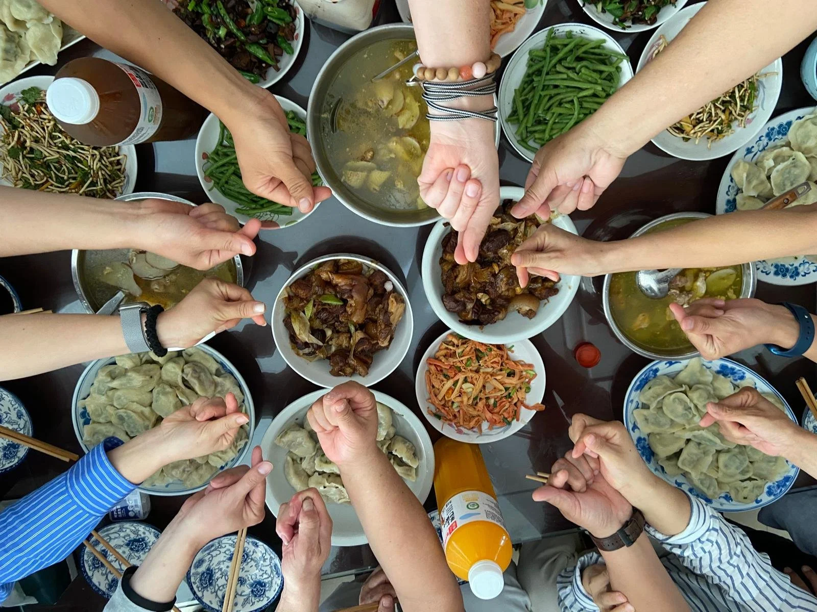 Overhead view of a table filled with various Asian dishes including dumplings, stir-fried vegetables, and broths, with multiple hands reaching in to share the meal.