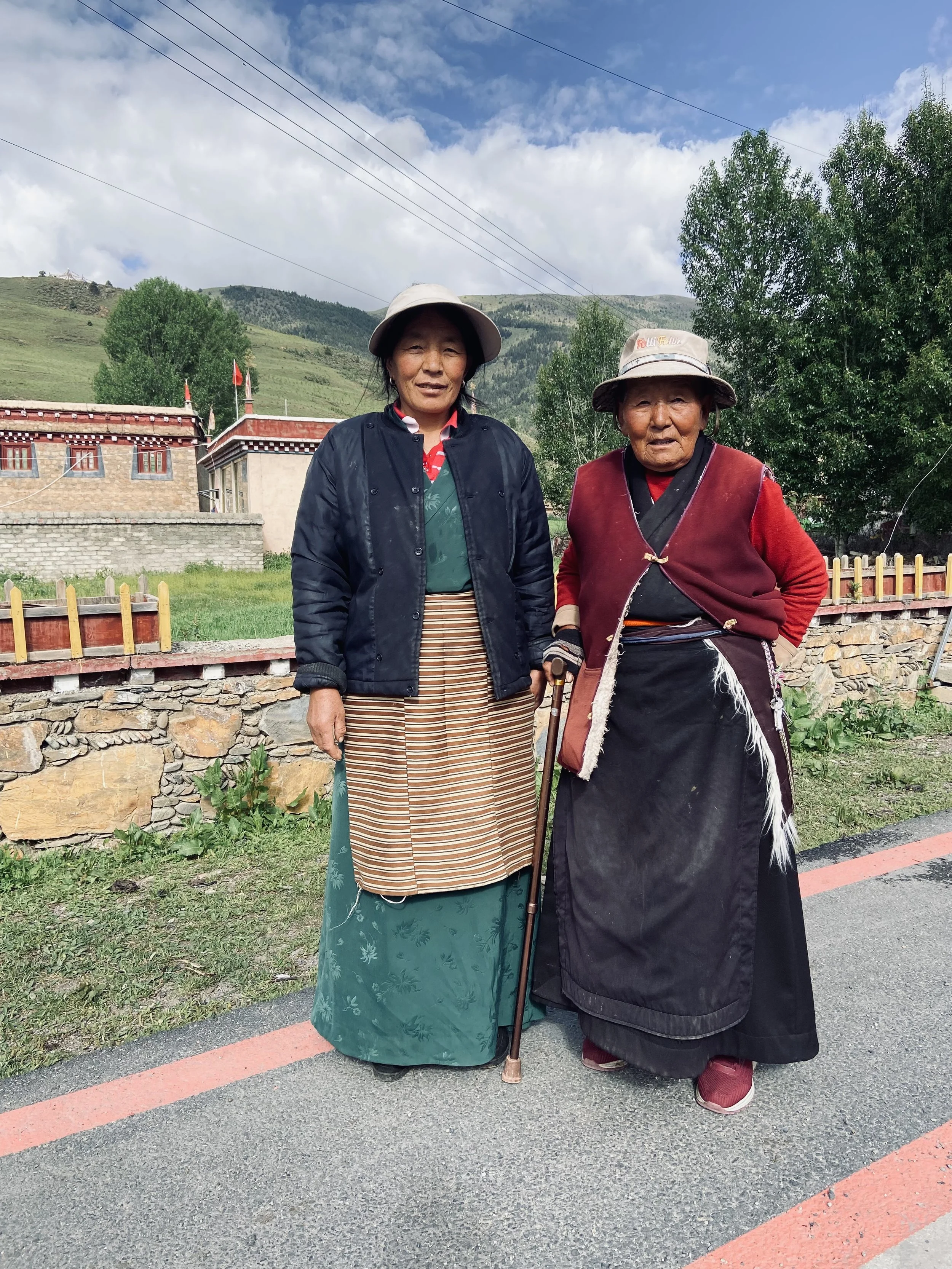 Two women in traditional attire standing outdoors in a rural area with green hills and a building in the background.