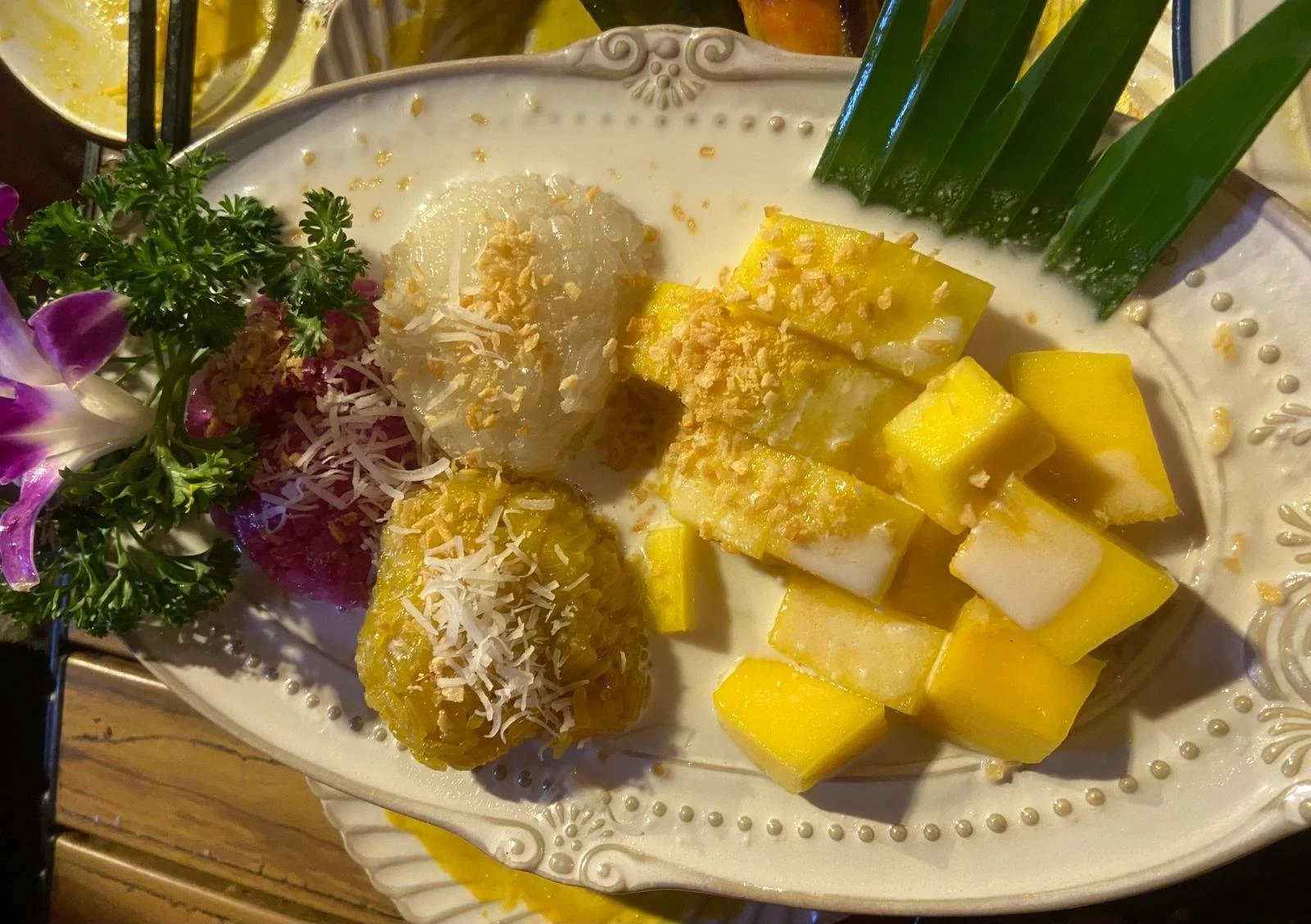 A plate of Thai mango sticky rice with fresh mango chunks, coconut sticky rice balls topped with shredded coconut, and garnished with parsley, purple orchids, and green leaves.