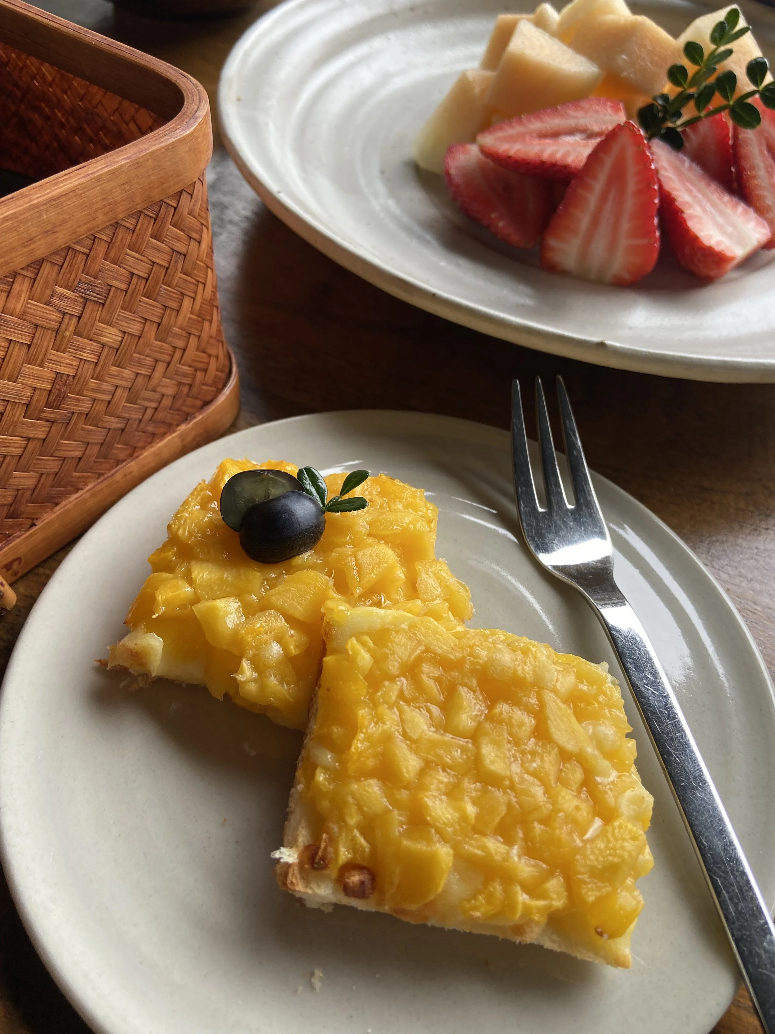 Slices of a yellow fruit dessert topped with two black cherries on a small white plate with a fork, with a bowl of sliced strawberries and melons in the background.