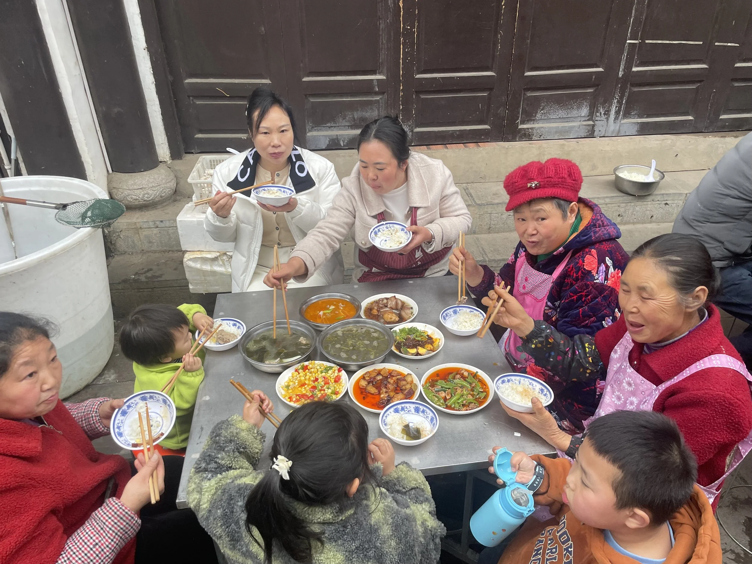 A group of people, including children and elderly women, sitting around a table outdoors, enjoying a meal with multiple bowls of soup, vegetables, and rice.