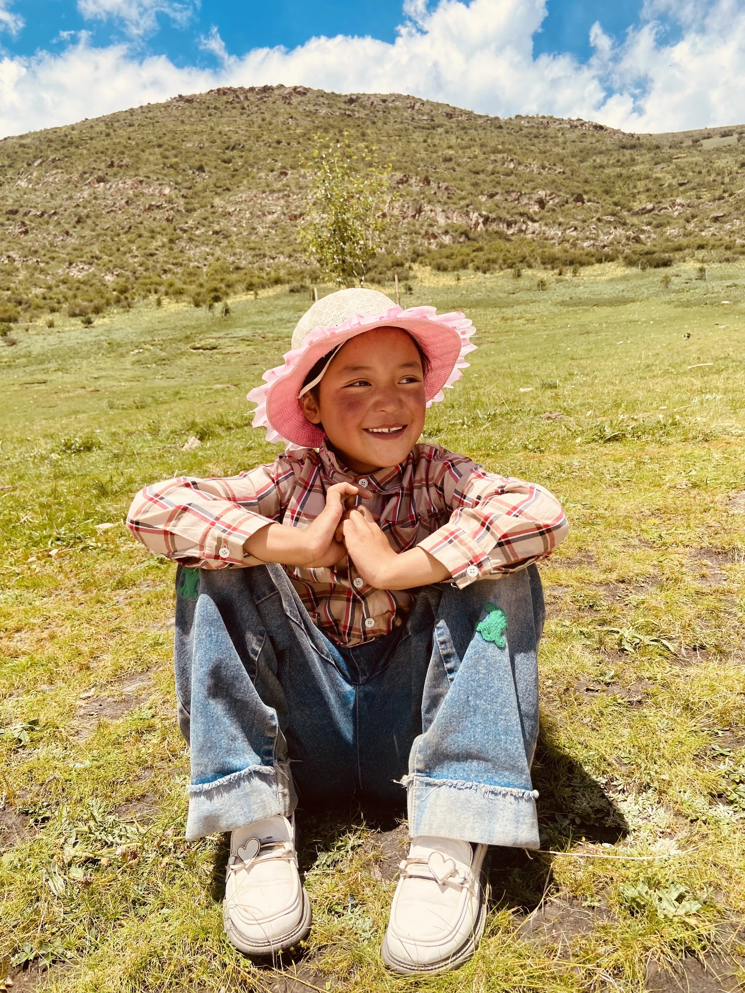 A young girl sitting on the grass in a field with mountains in the background. She is wearing a large pink and white sun hat, a plaid shirt, ripped jeans, and white shoes, smiling happily.